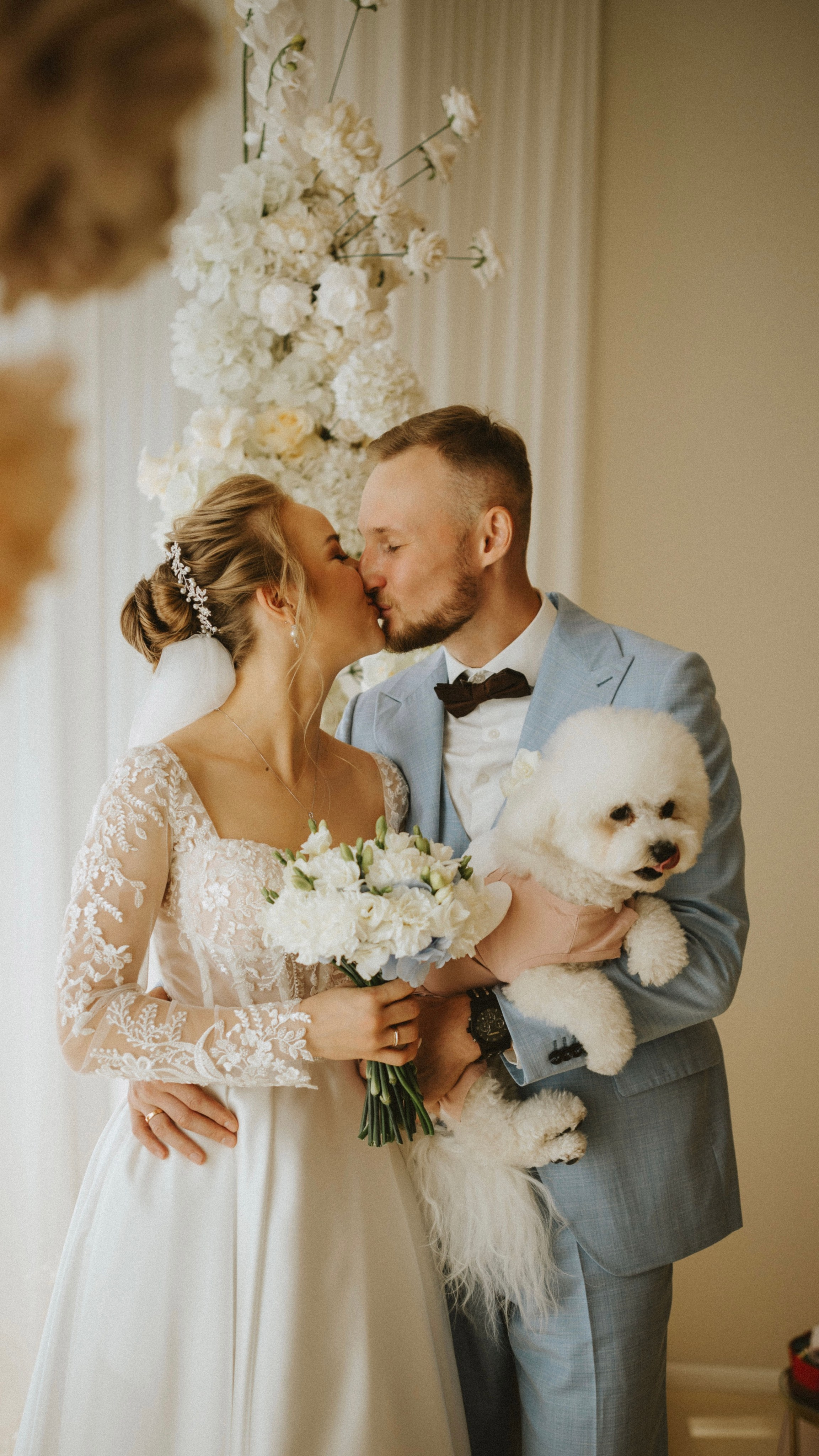 “Una sposa sta serenamente in un momento di calma, con in mano un bouquet di piccoli fiori bianchi. Ha un'espressione pacifica sul viso, con gli occhi dolcemente chiusi. Il suo abito da sposa presenta delicati dettagli in pizzo e indossa un bellissimo diadema. La luce soffusa mette in risalto i suoi lineamenti, creando un'atmosfera serena. La parete bianca ornata alle sue spalle completa l'atmosfera elegante del momento”.