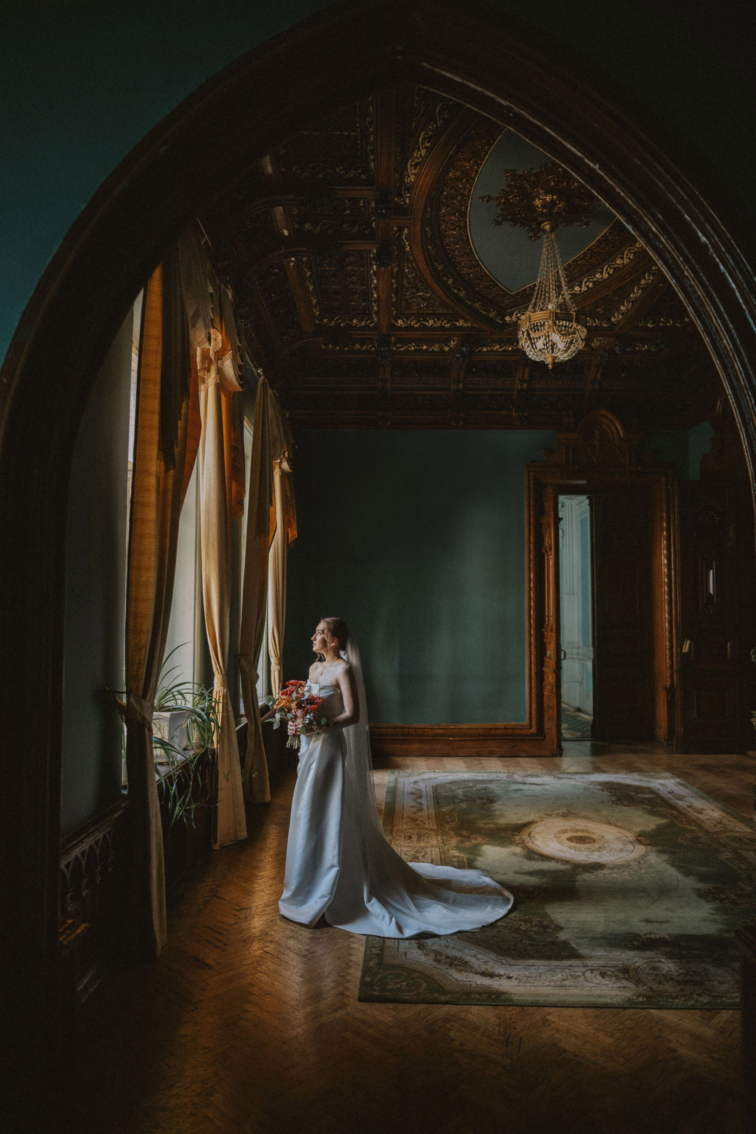 “Una sposa in abito senza spalline si affaccia con grazia alla grande finestra di una stanza elegante e decorata. Con una mano tiene un bouquet di fiori, mentre con l'altra tocca delicatamente la cornice della finestra. La stanza presenta un soffitto intricato, rifiniture in legno e pareti di un bel verde intenso, con una luce morbida e calda che entra dalla finestra”.