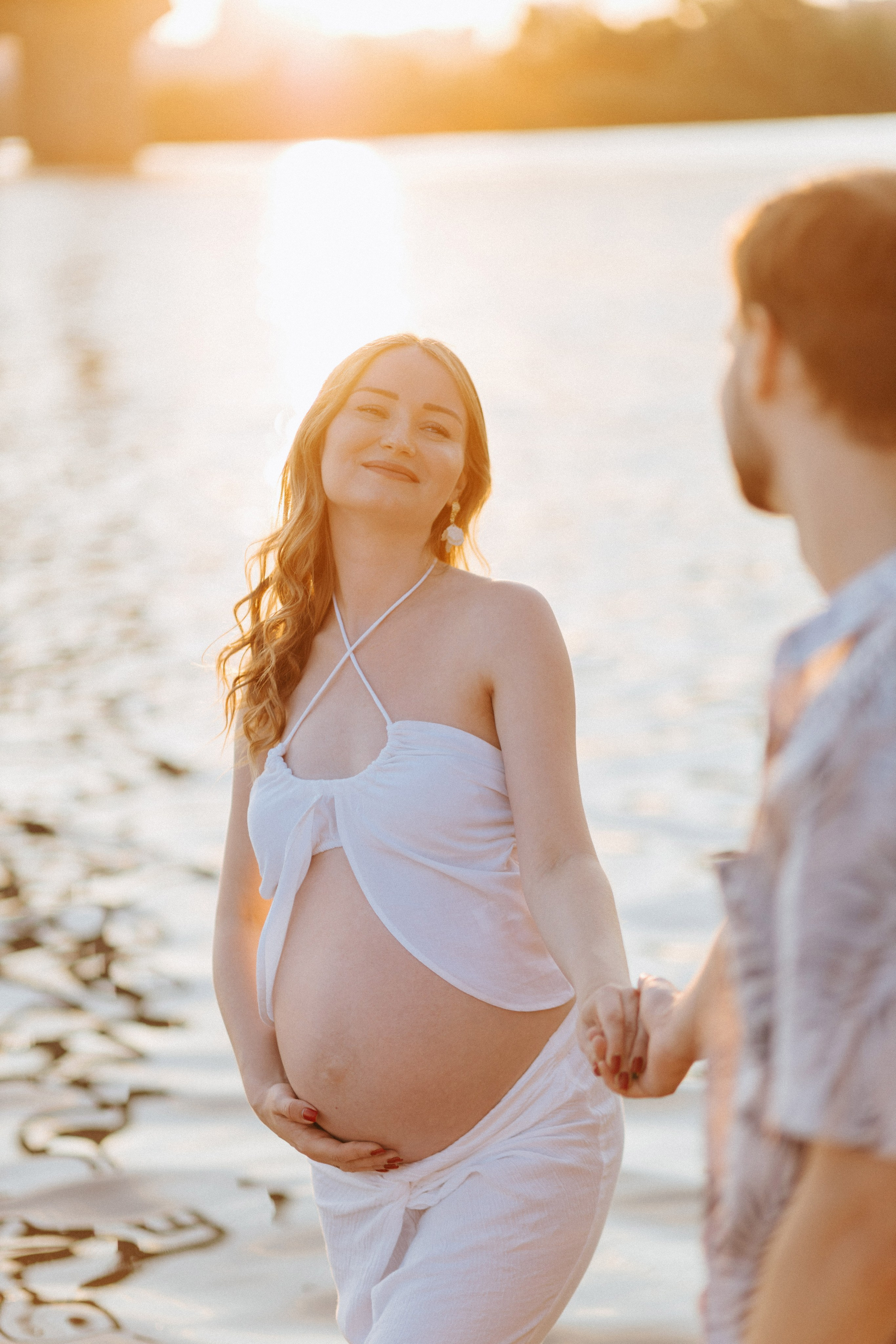“Una luminosa donna incinta in abito bianco, che cammina lungo l'acqua tenendo per mano il suo compagno. La luce dorata del tramonto proietta un caldo bagliore sul suo viso, mettendo in risalto la sua espressione gioiosa. L'acqua calma riflette le tonalità calde della sera, aggiungendo un'atmosfera serena a questo momento intimo”.