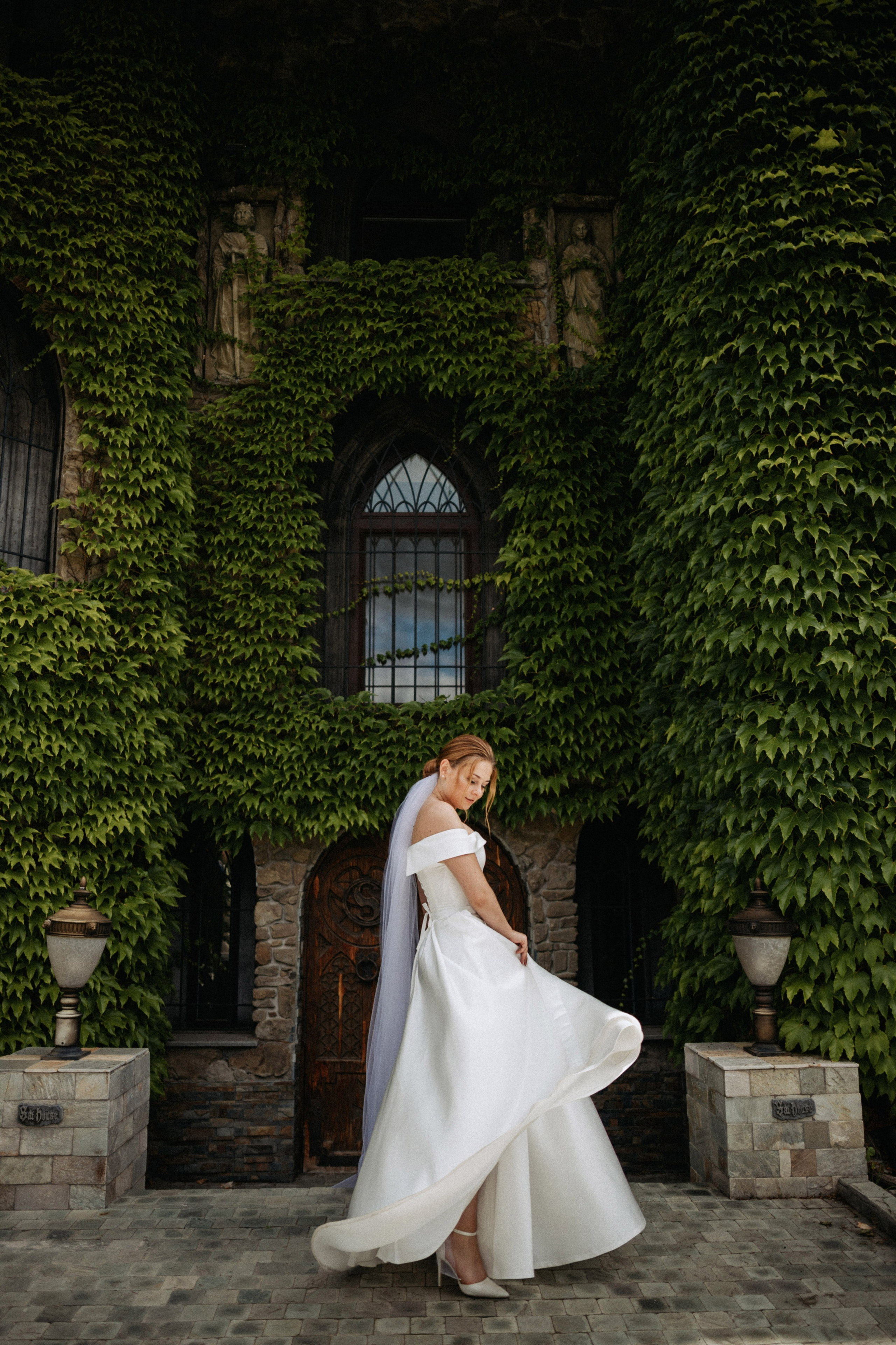 “La sposa si erge con grazia davanti a un bellissimo edificio ricoperto di edera, con il suo abito da sposa che scorre elegantemente mentre guarda verso il basso. Il morbido stile off-the-shoulder del suo abito e il suo velo aggiungono un'atmosfera romantica, rendendo questo momento perfetto catturato in un ambiente naturale”.