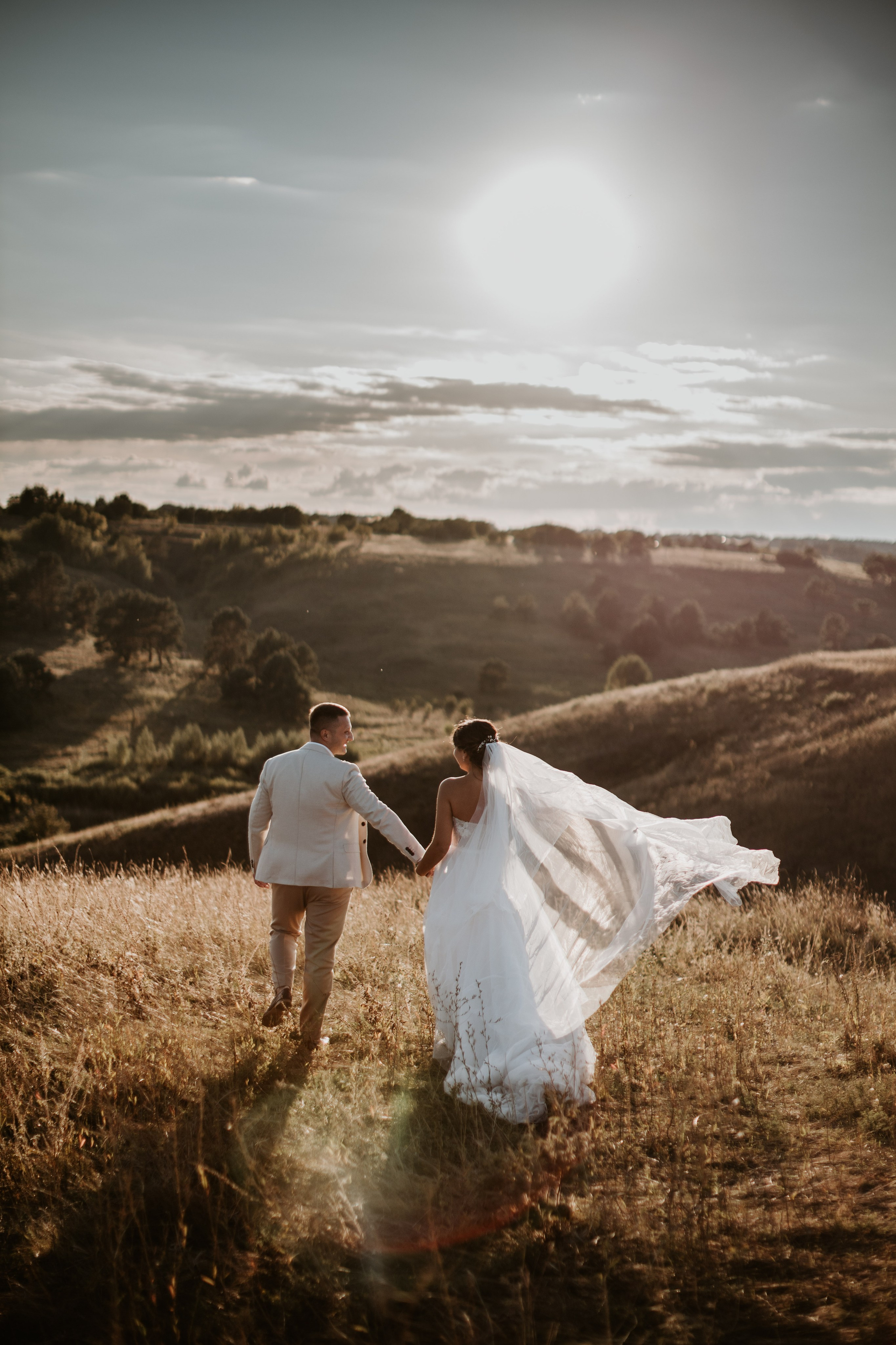 “Una coppia di sposi che cammina mano nella mano su una collina erbosa durante il tramonto, con il velo della sposa che scorre nel vento. La morbida luce dorata del sole illumina la coppia, creando un'atmosfera romantica e serena mentre si godono il loro momento speciale circondati dalla natura.”