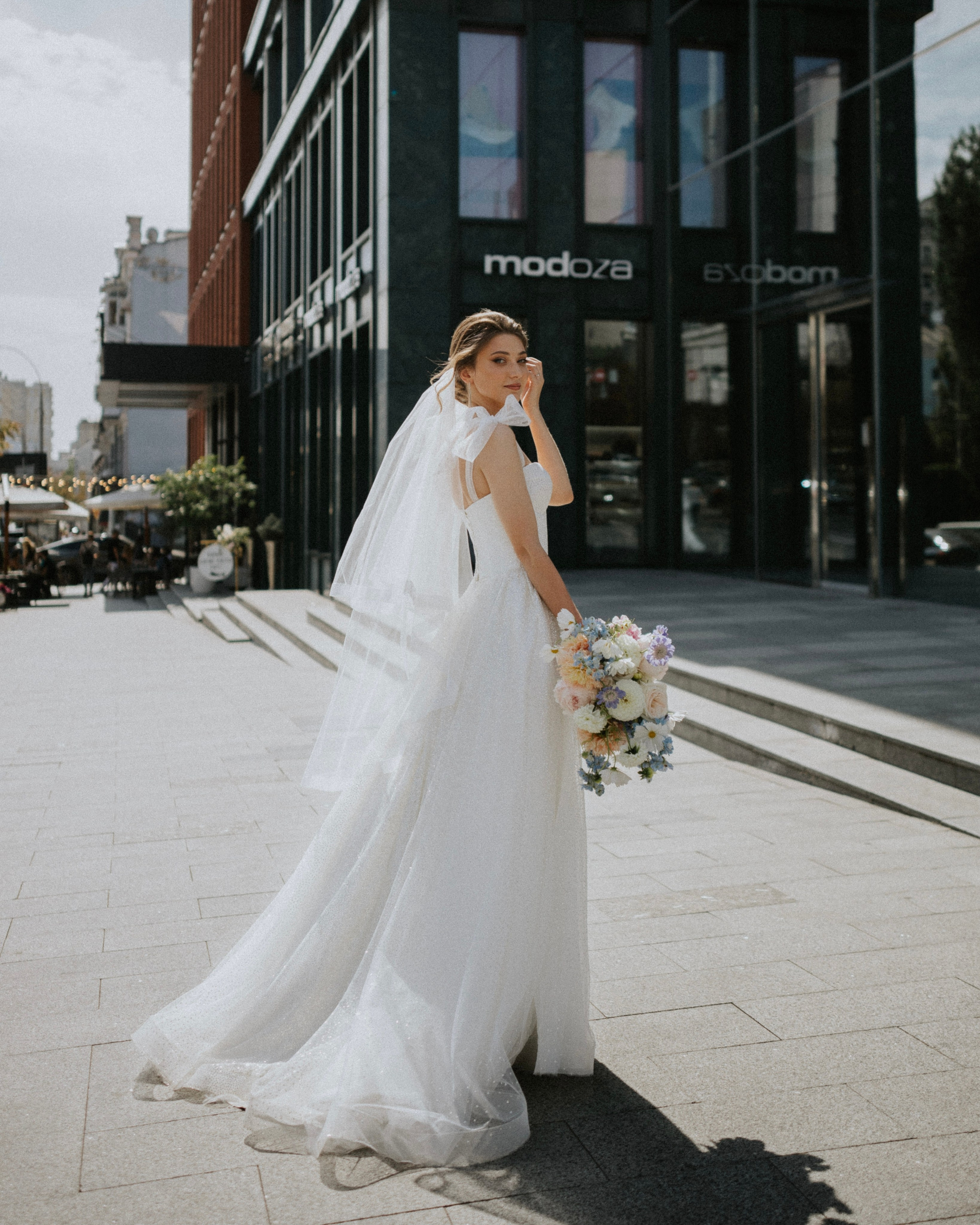 “Fotografo di matrimonio che immortala una splendida sposa in un fluente abito bianco, con in mano un bellissimo bouquet di fiori pastello, mentre cammina in una moderna strada cittadina. La sposa, con un velo delicato e un sorriso tenero, irradia eleganza, rendendo questo momento perfetto per un matrimonio urbano in Italia.”