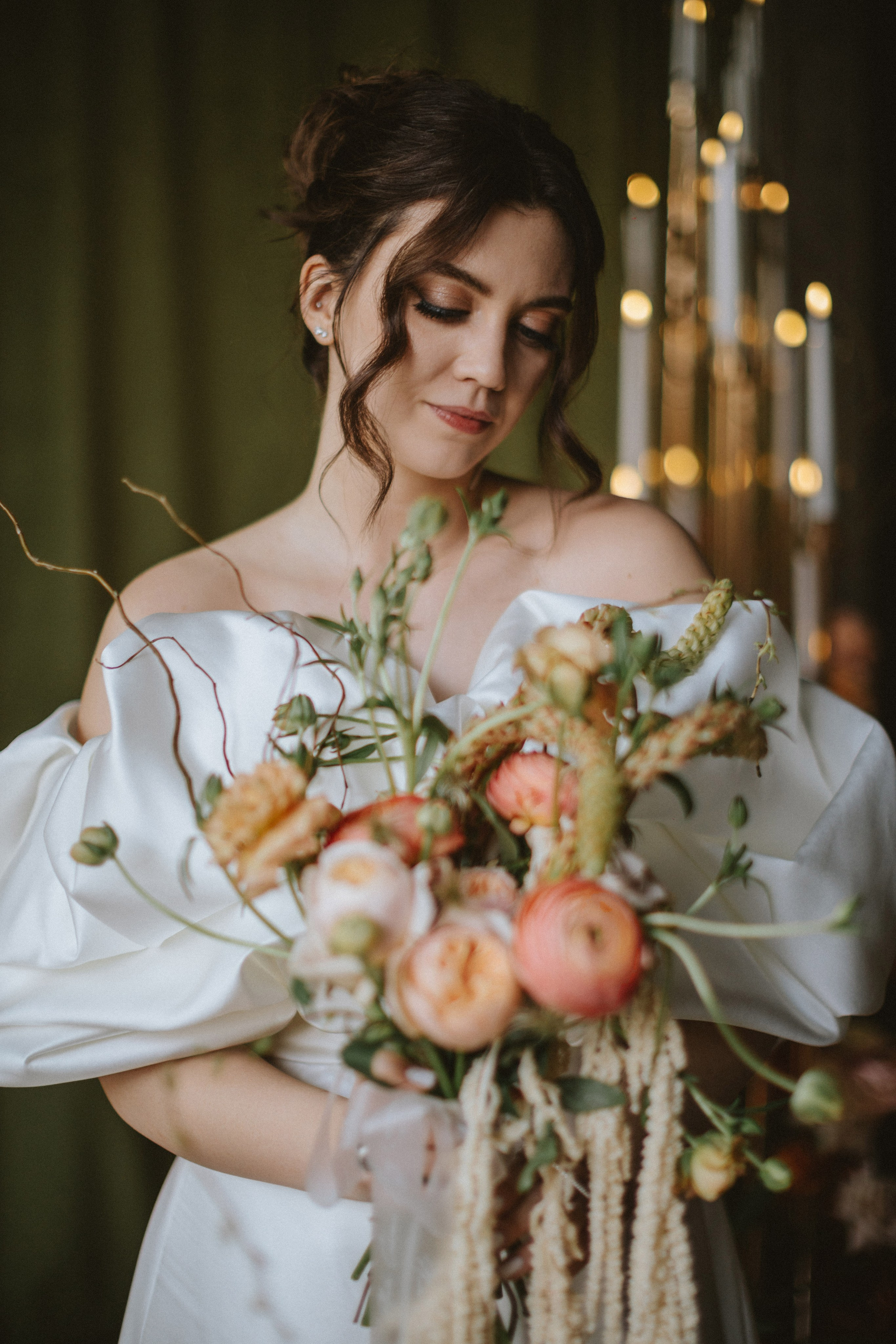 “Una sposa tiene delicatamente in mano un lussureggiante bouquet di peonie rosa tenue, ranuncoli delicati e vegetazione in pendant. Il suo abito da sposa off-the-shoulder, con drammatiche maniche a sbuffo, incornicia il suo look elegante mentre guarda dolcemente verso il basso, circondata da un ambiente caldo di candele luminose sullo sfondo”.