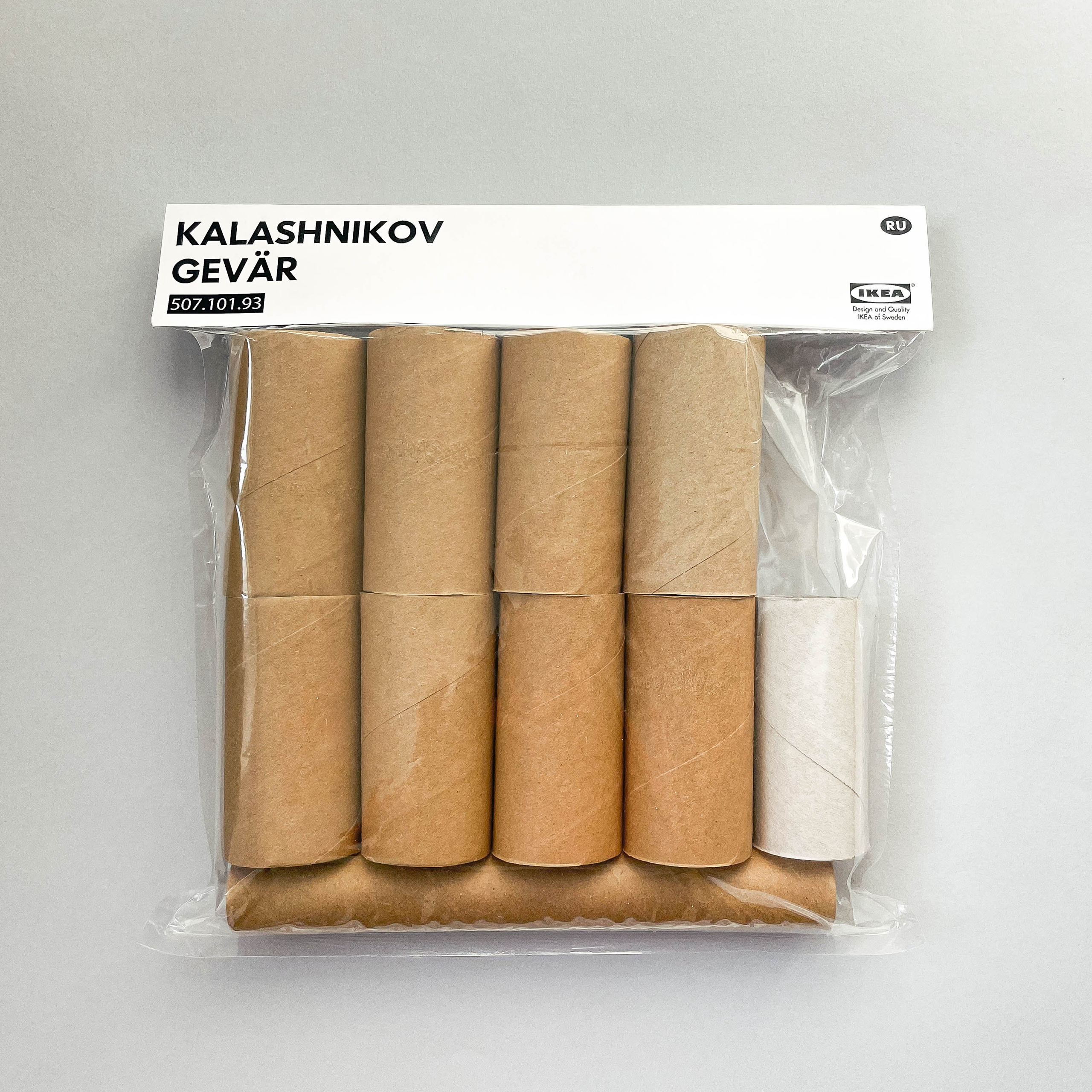 The image displays a package of toilet paper rolls designed to resemble an IKEA product. The packaging is labeled "KALASHNIKOV GEVÄR," which translates to "Kalashnikov Rifle" in Swedish. The design mimics typical IKEA packaging, with a white label at the top, including a product code (507.101.93) and an IKEA logo. The package contains several brown cardboard toilet paper rolls, arranged neatly in two rows, with one white roll in the bottom right corner. The background is a plain, light gray surface.