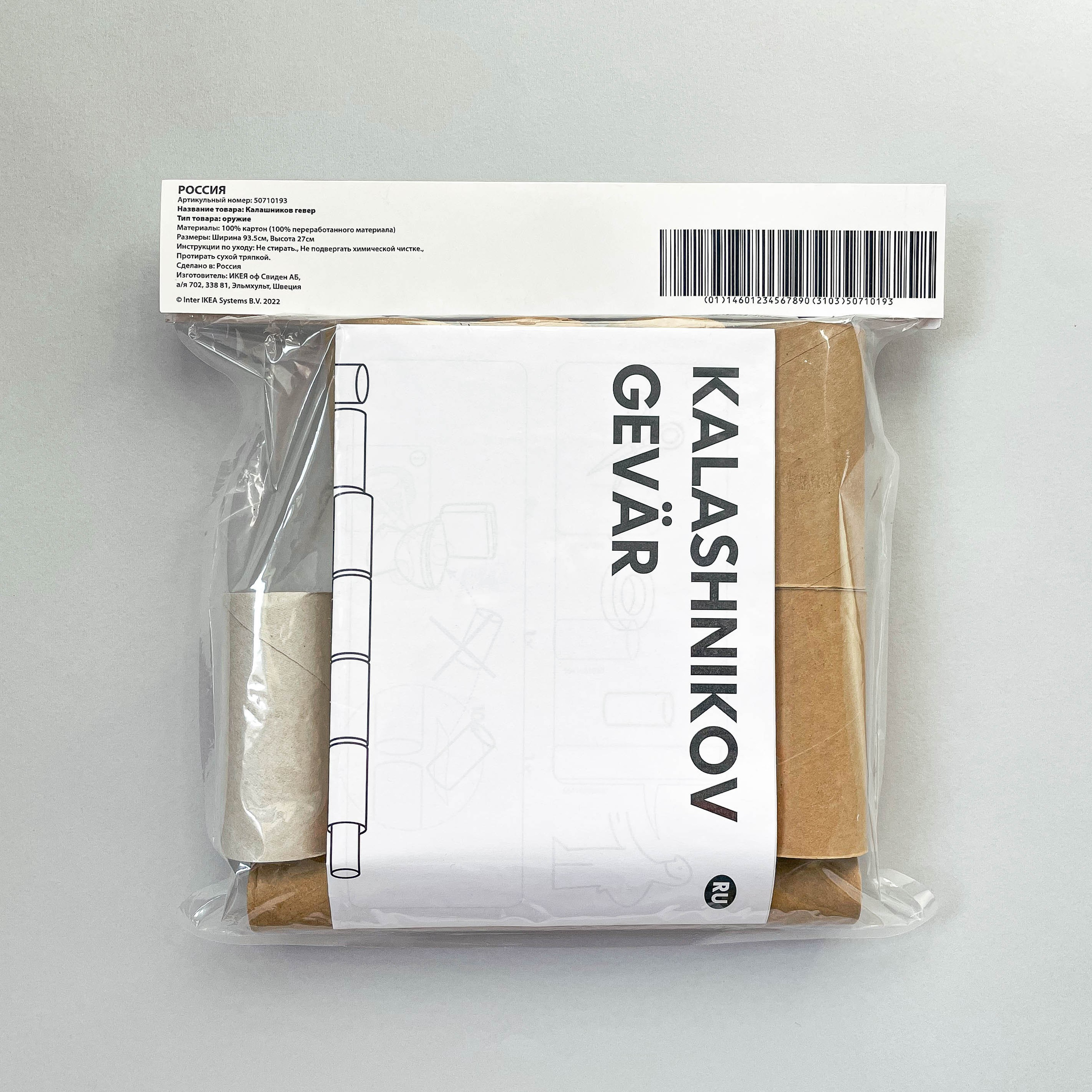 The image shows the backside of a package of toilet paper rolls designed to look like an IKEA product. The package label reads "KALASHNIKOV GEVÄR," which translates to "Kalashnikov Rifle" in Swedish. The label includes a barcode and product information in Russian, detailing the item as an environmentally friendly product made from recycled materials. The rolls are visible through the transparent plastic packaging. The background is a plain, light gray surface.
