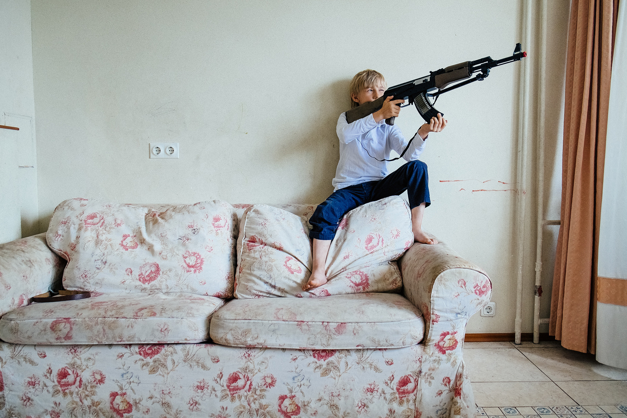 A boy with a gun is sitting on the couch. Мальчик с ружьем сидит на диване.