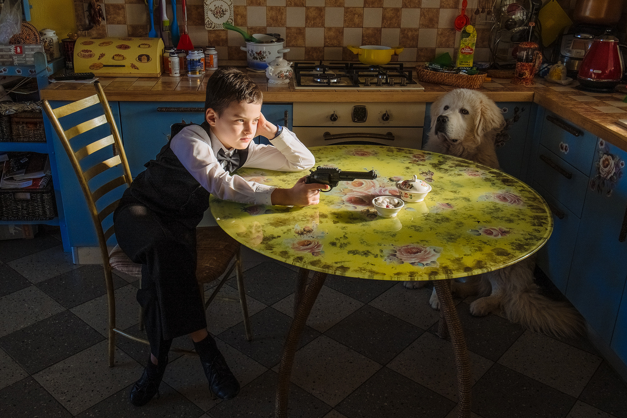 A boy is sitting at a table and holding a gun, and a big white dog is sitting next to him. Мальчик сидит за столом и держит в руках пистолет, а рядом сидит большая белая собака.
