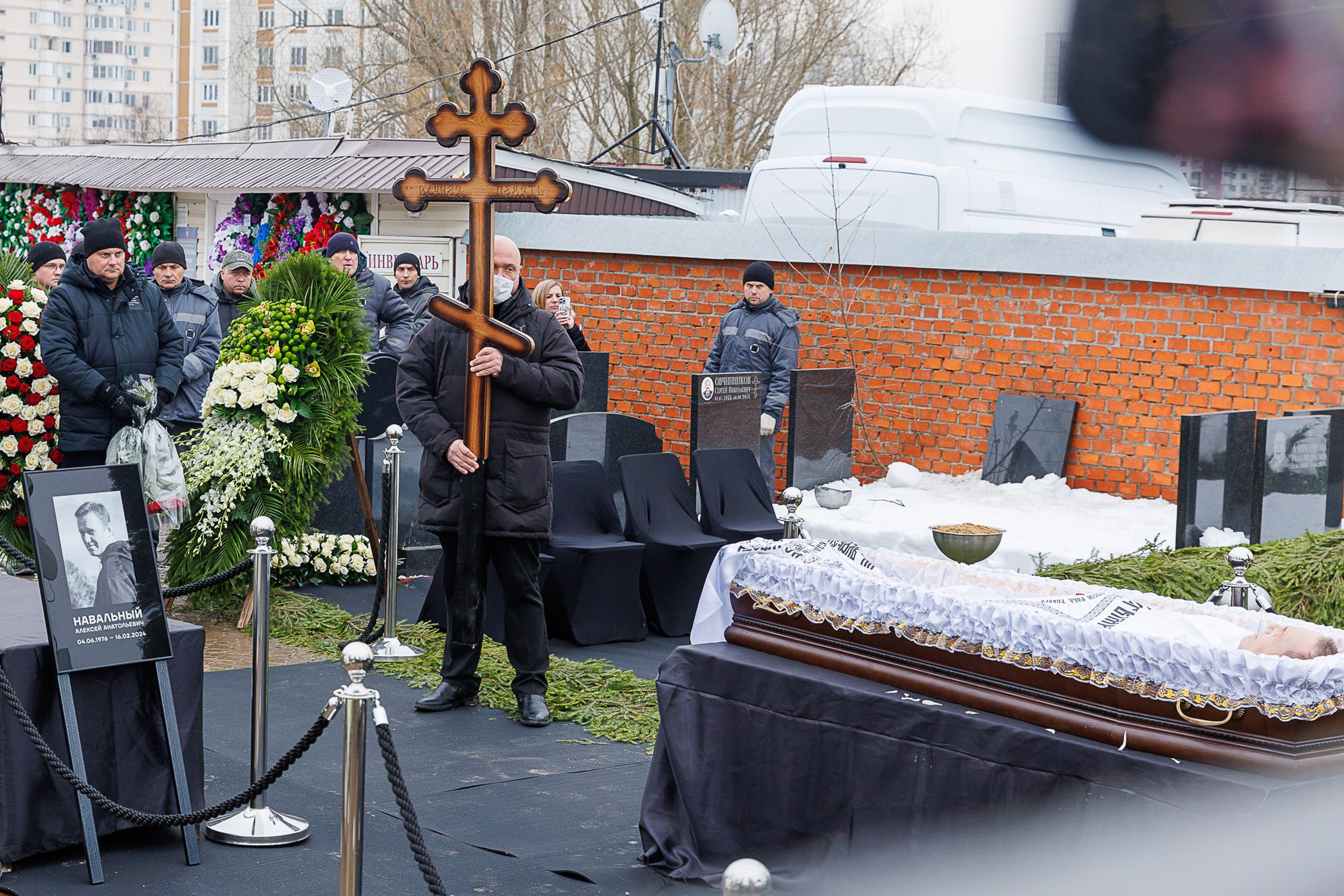 Navalny lying in coffin surrounded by flowers and priest, 2024
