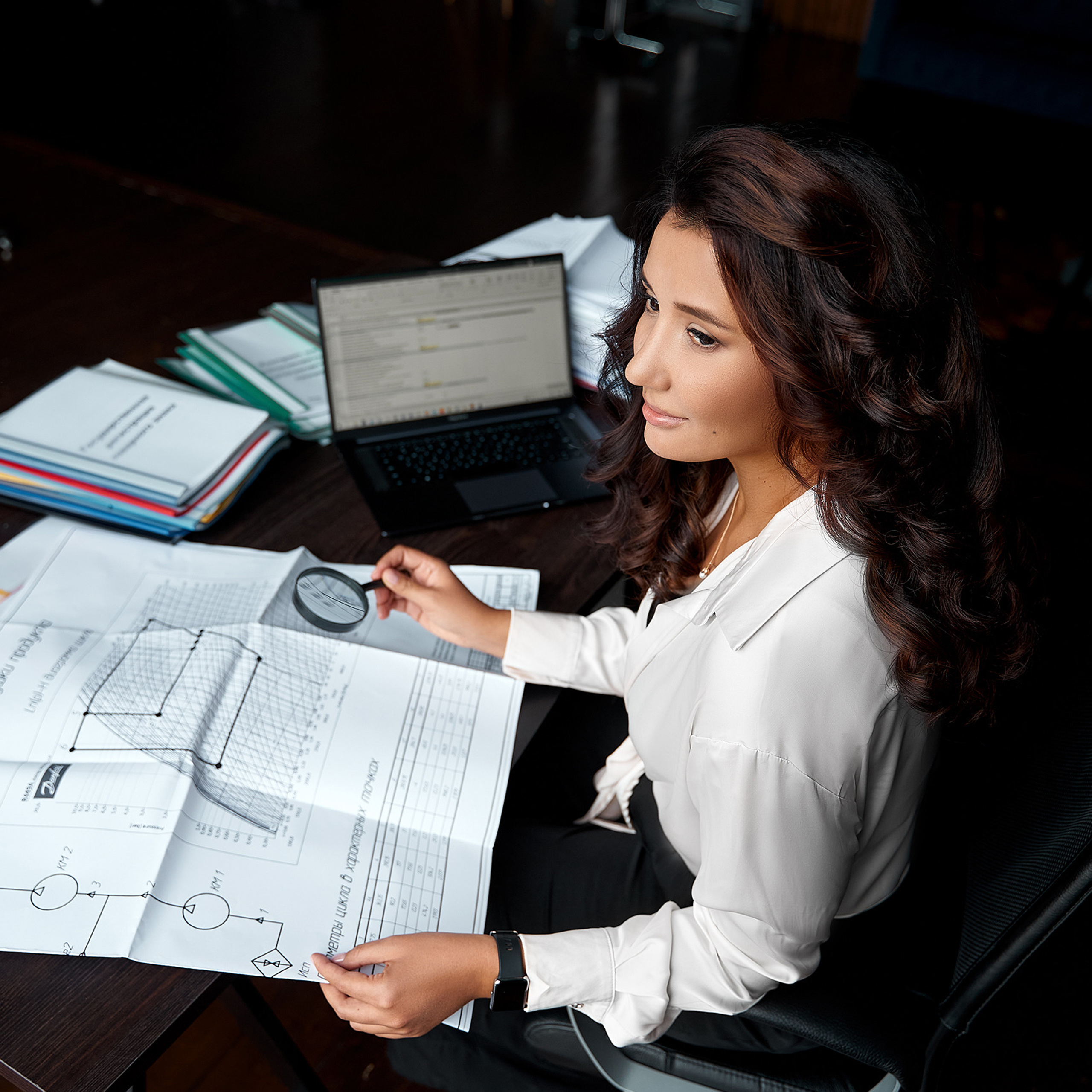 Professional female analyst examining complex charts and graphs with a magnifying glass in a well-organized office space.