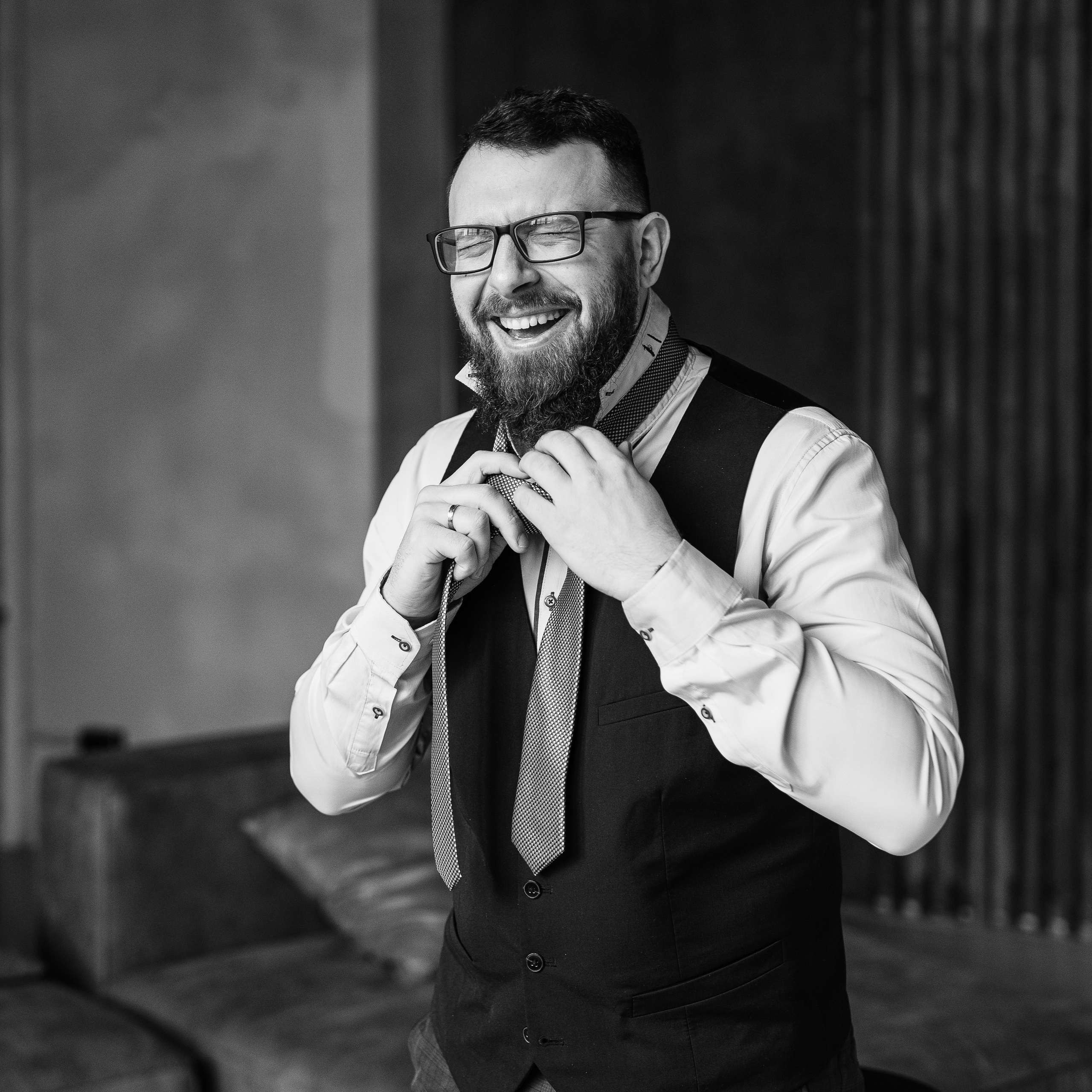 Smiling bearded man adjusting his tie in a black vest and white shirt, black and white photo