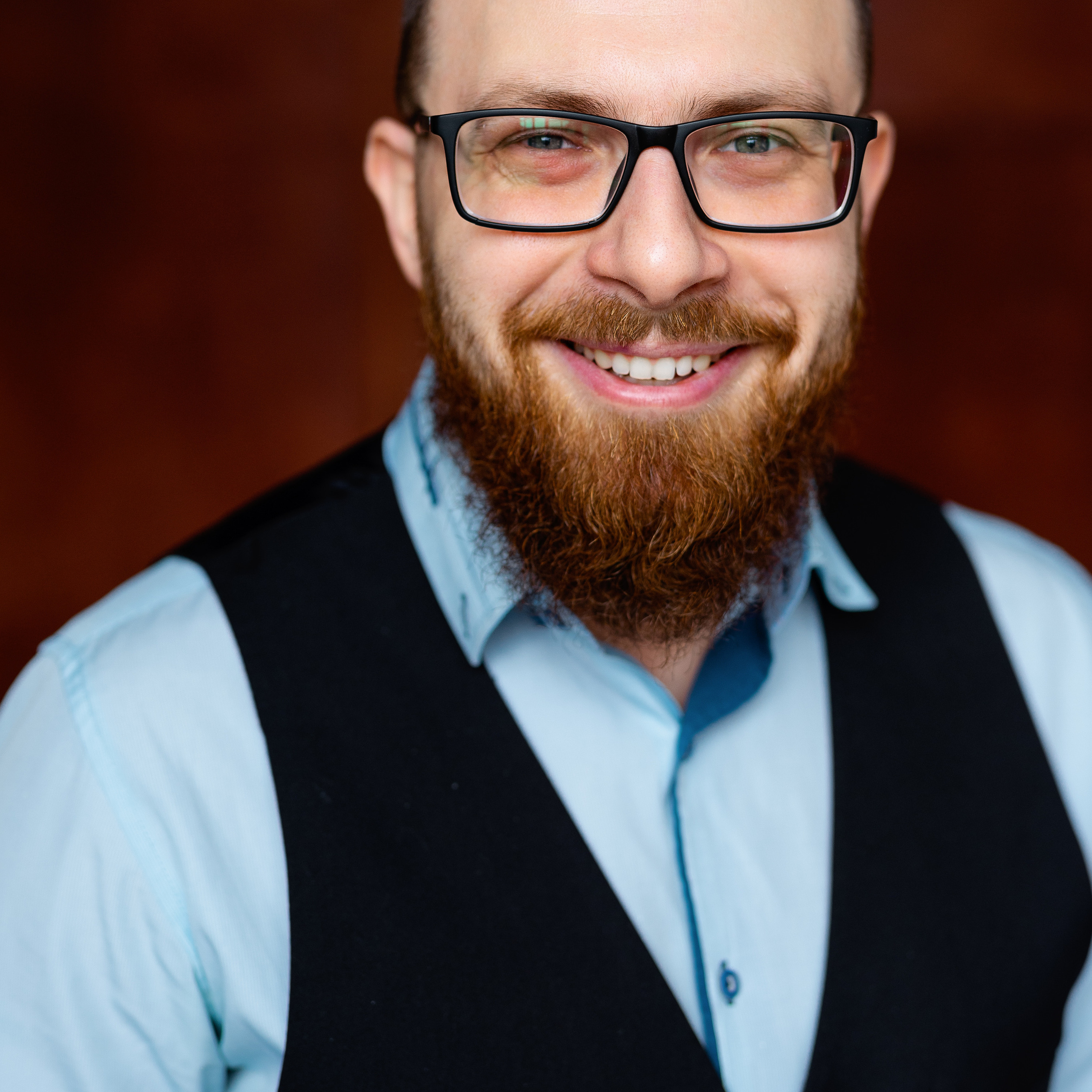 Portrait of a cheerful bearded man with glasses wearing a light blue shirt and dark vest, conveying a friendly and professional demeanor.
