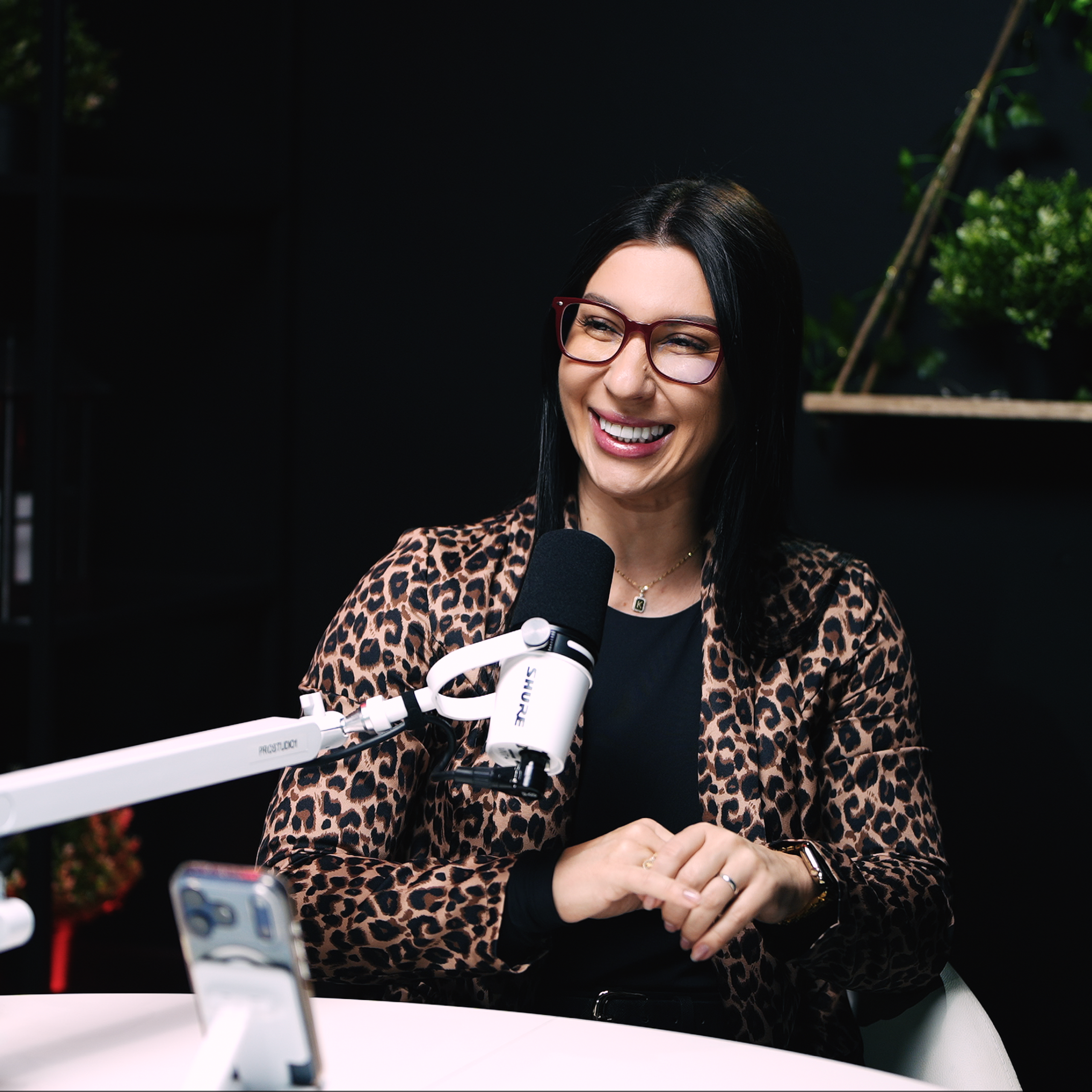 Podcast guest on mic at ProStudio1, Hollywood FL — seated interview shot with cinematic dark background and greenery accents.
