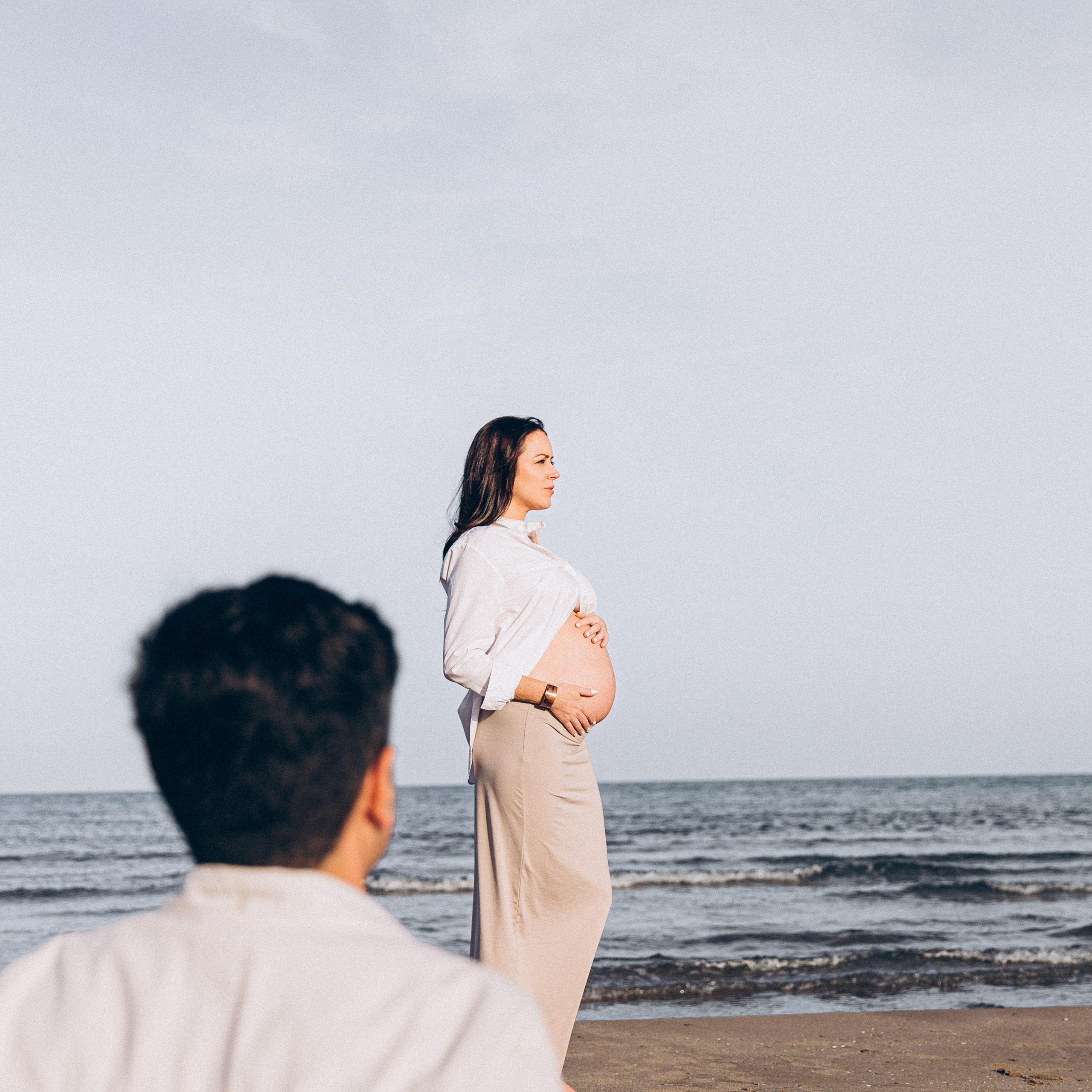 Maternity photoshoot on beach with pregnant woman in white blouse and beige pants gazing at horizon, man in foreground, in Valencia, Spain.