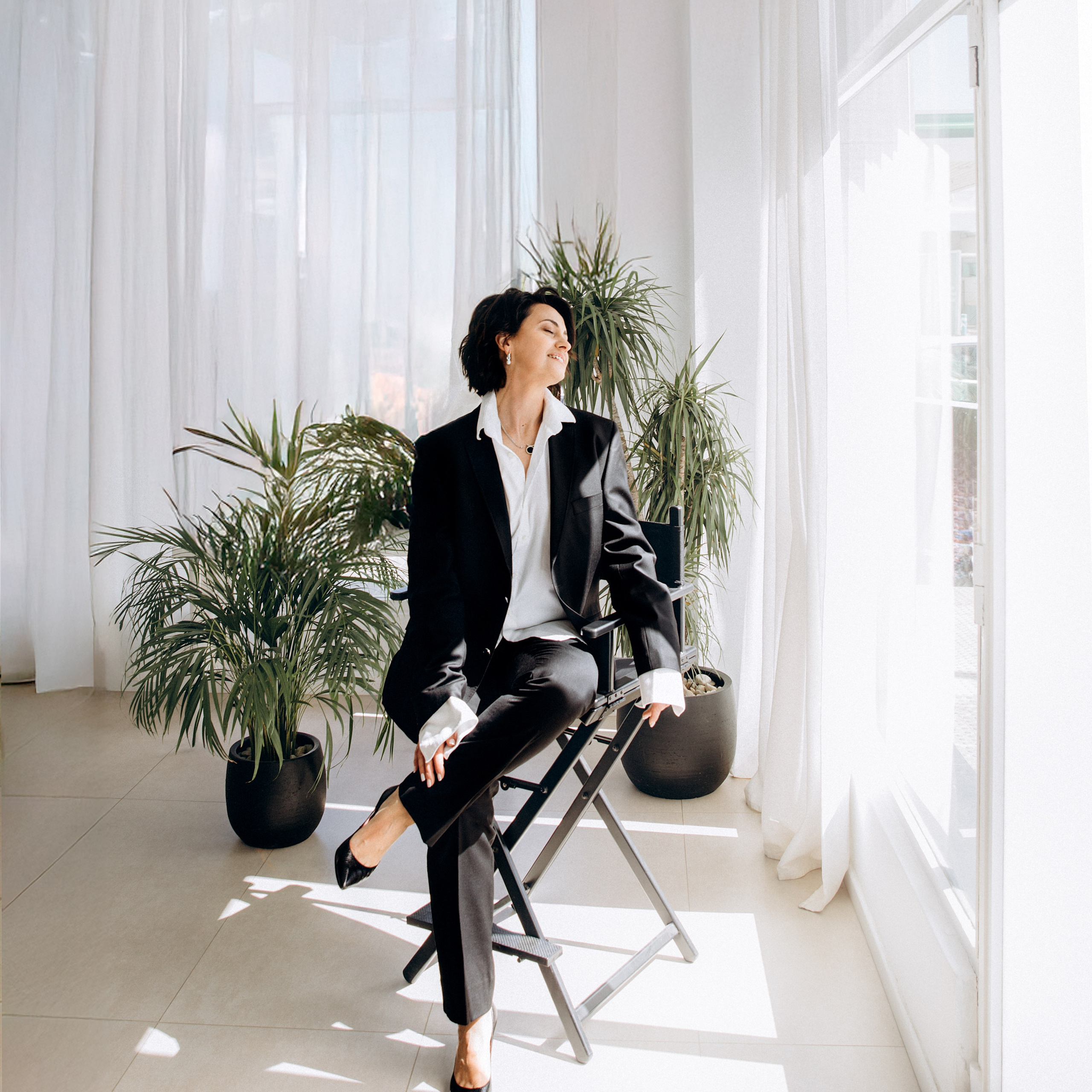 Professional portrait of a woman in a black suit sitting on a director’s chair surrounded by tropical plants and sunlight in a modern studio in Valencia, Spain