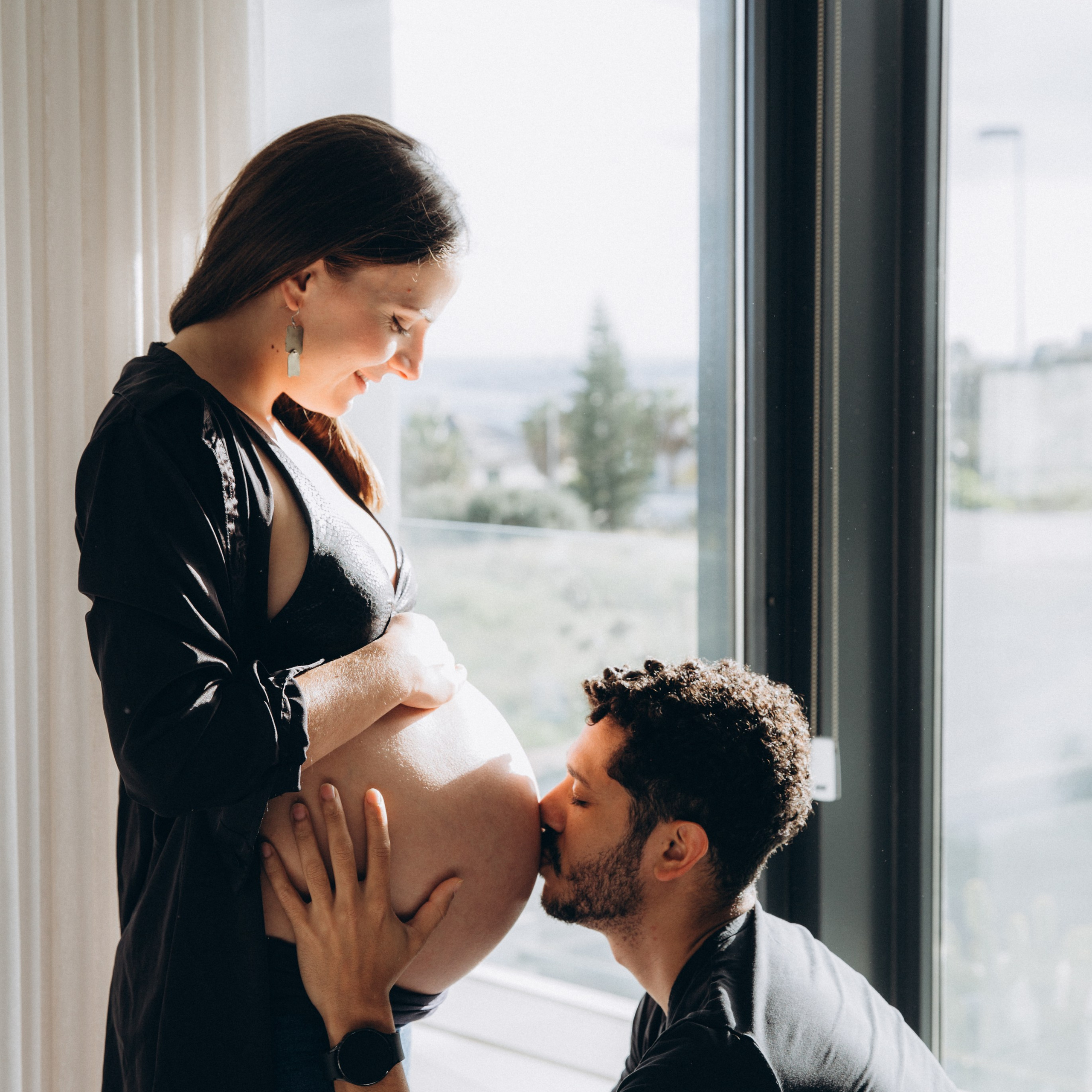 Intimate maternity photoshoot with a man kissing pregnant woman’s belly in a sunlit room, overlooking the cityscape, in Valencia, Spain