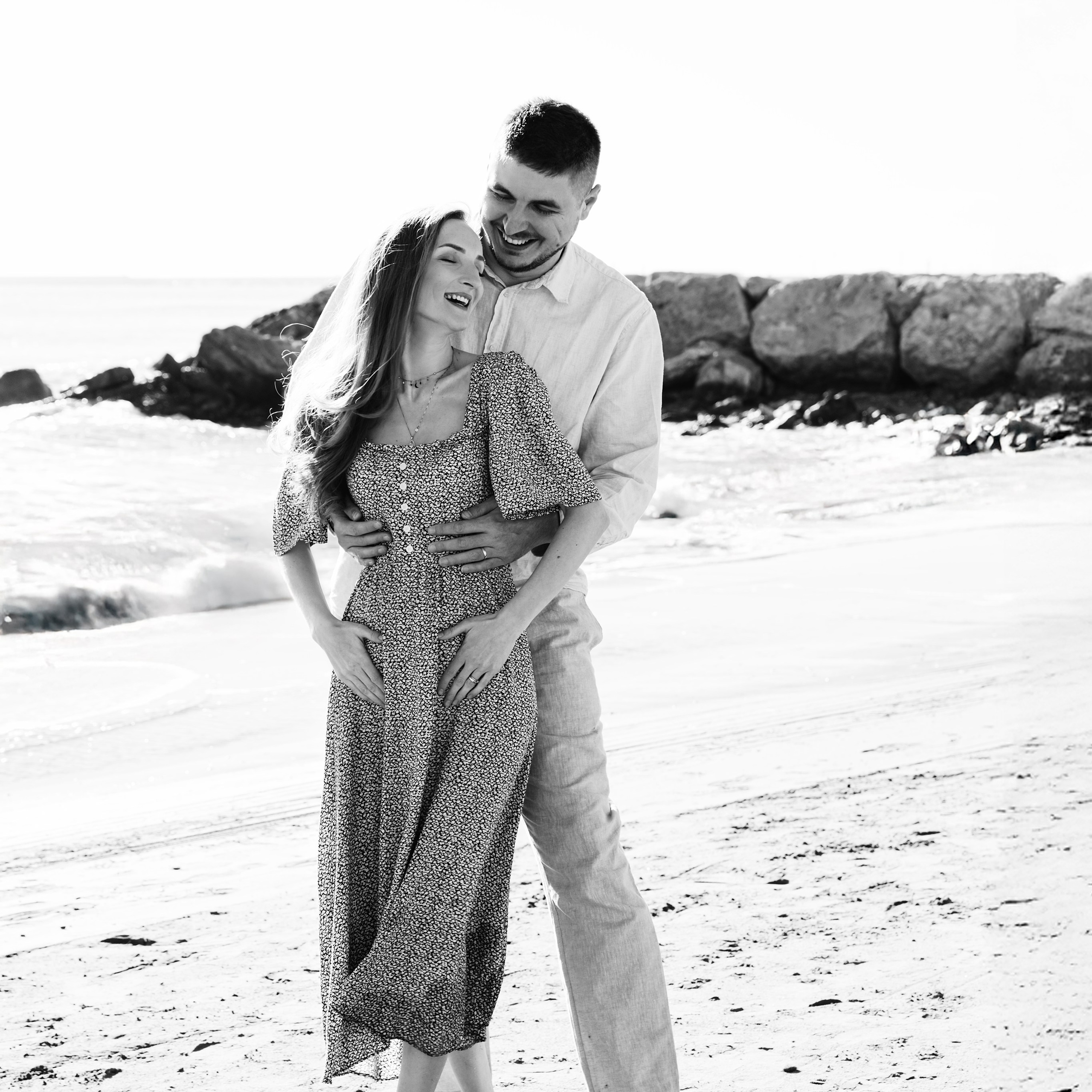 Joyful black and white couple’s photoshoot (love story) on the beach with a man embracing a smiling woman, barefoot on the sand, in Valencia, Spain