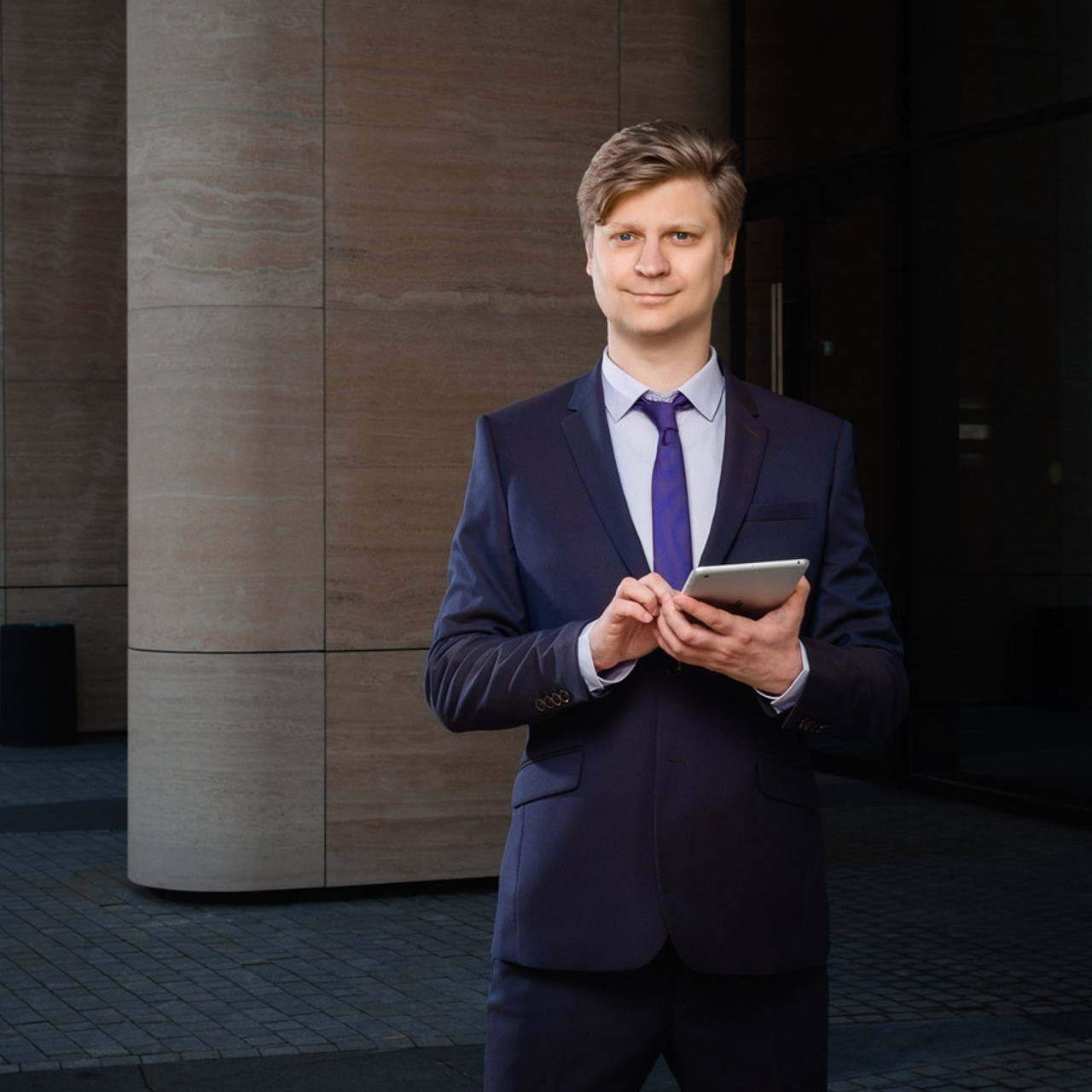 Businessman in a navy blue suit holding a tablet outside a modern building, a corporate headshot by Dmitrii Shmatov.