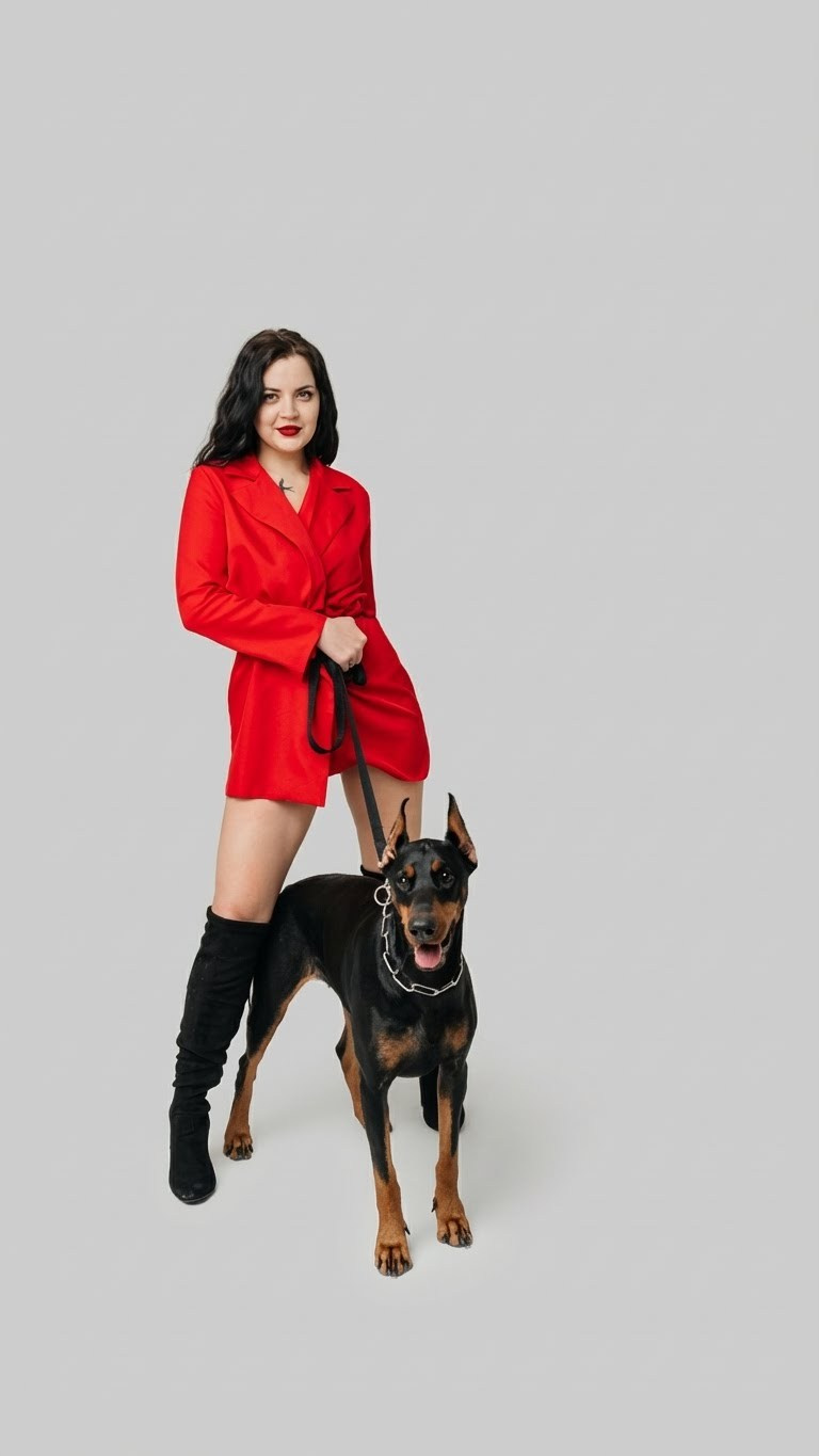 Dobermann springt neben einer Frau im roten Outfit vor hellem Hintergrund.