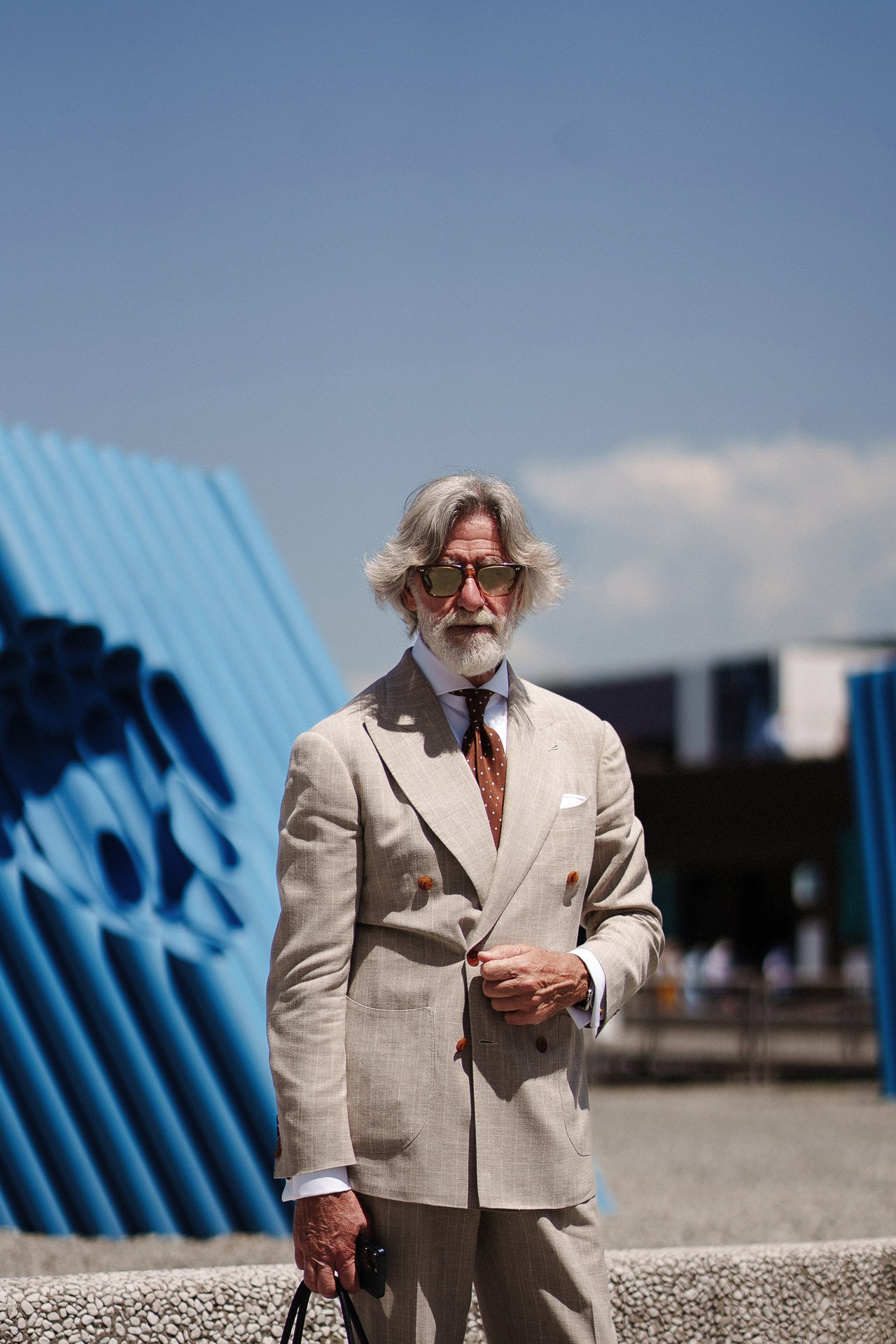 Ritratto street style da Pitti Uomo 110, uomo più adulto in completo doppiopetto beige vicino all’installazione blu