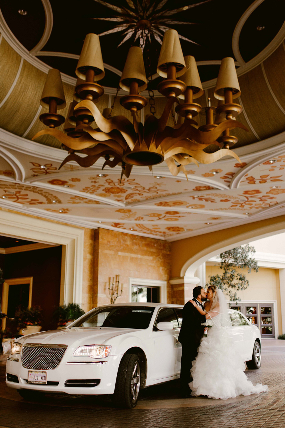 The bride and groom kiss in front of the wedding building in front of a white wedding limousine. Above their heads is a huge chandelier.