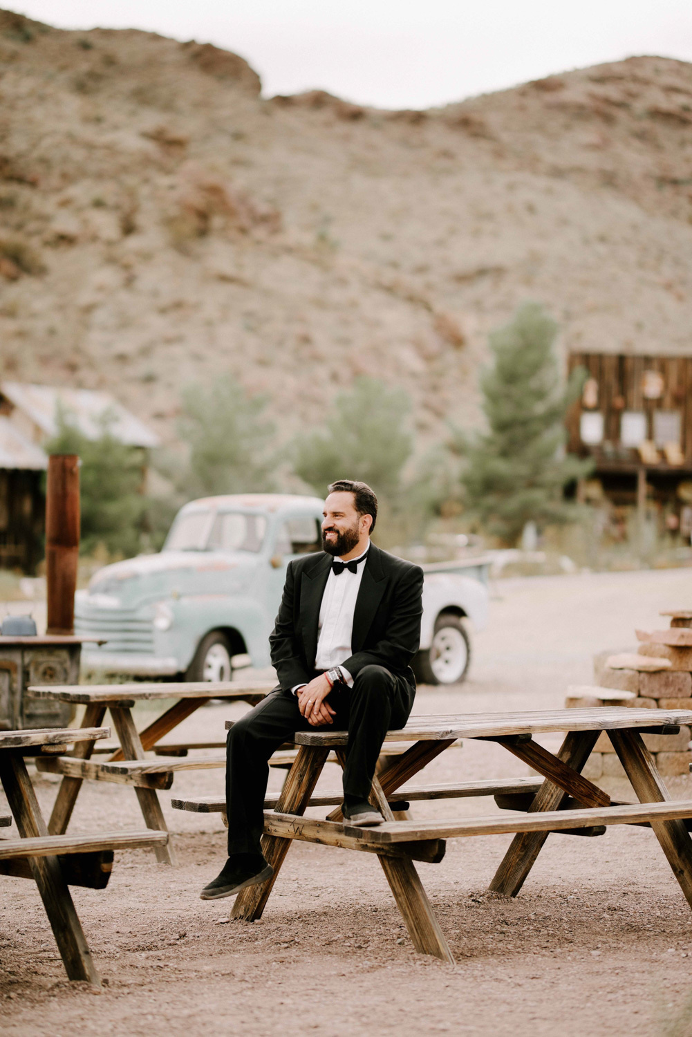 The groom is sitting on a wooden table with an old white car on the background of mountains and green bushes.