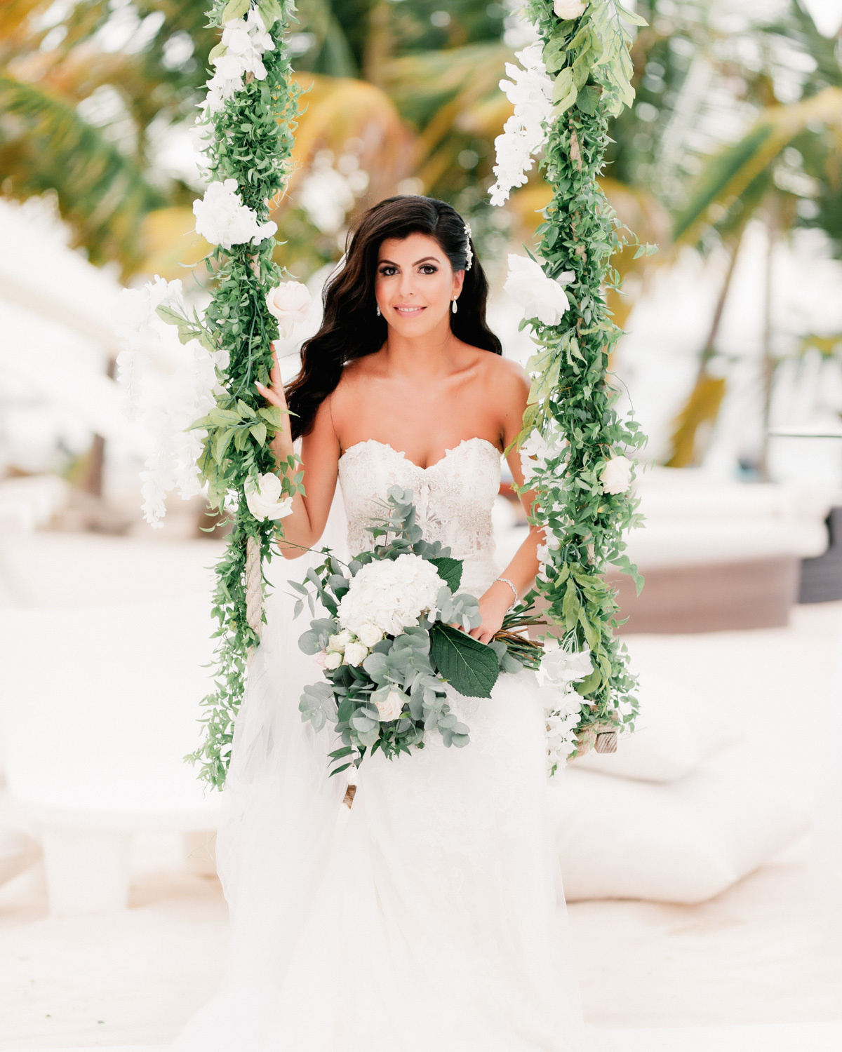 Dark-haired girl with loose hair in a white wedding dress with a bouquet of white flowers stands on the background of a sea beach with palm trees.