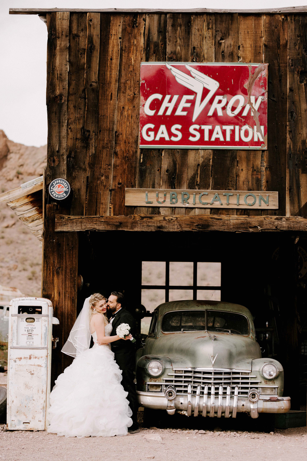 Bride and groom stand at an old abandoned gas station, with a vintage car in the background.