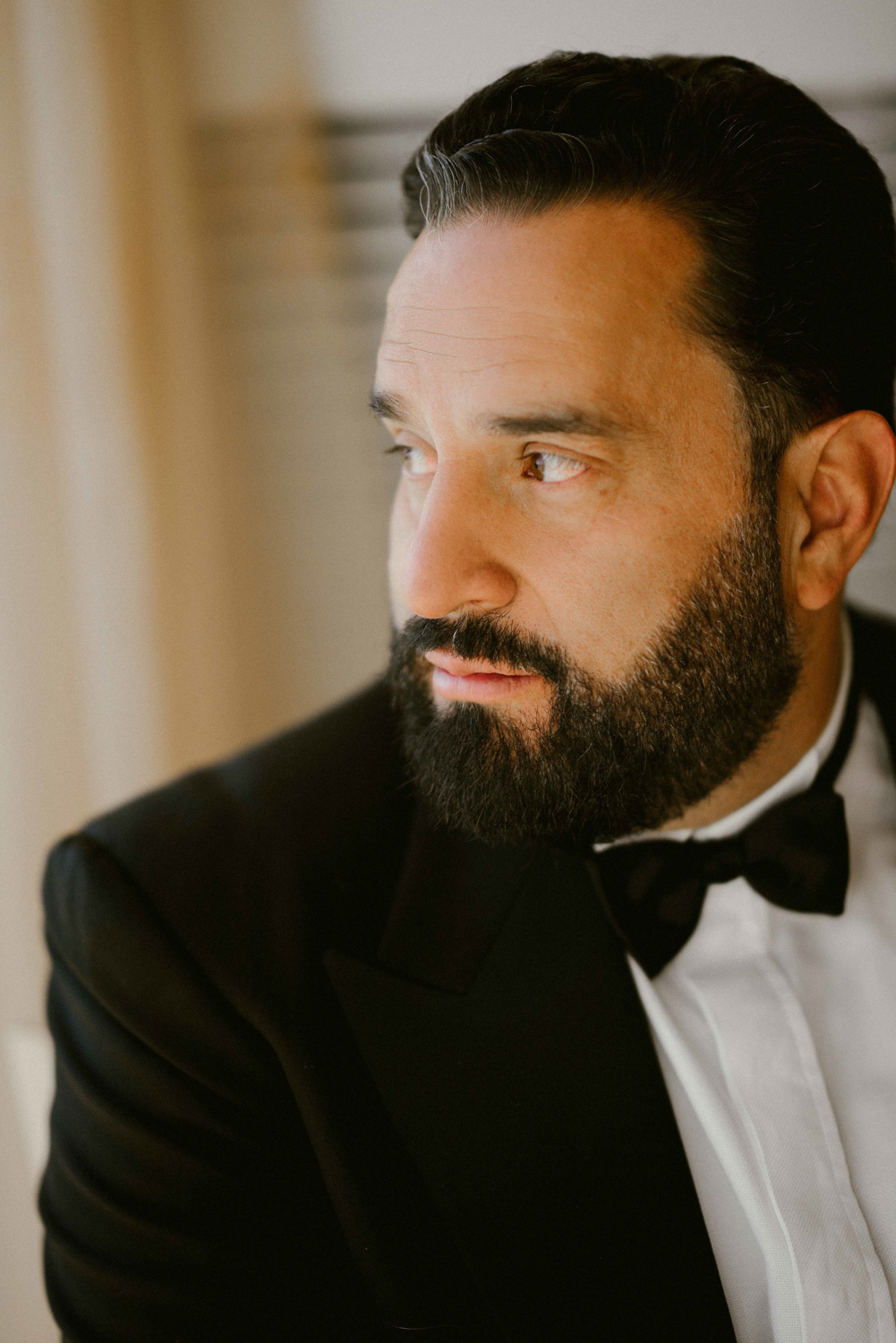 Black-haired and bearded groom in a dark suit with a dark bow tie on a white shirt.