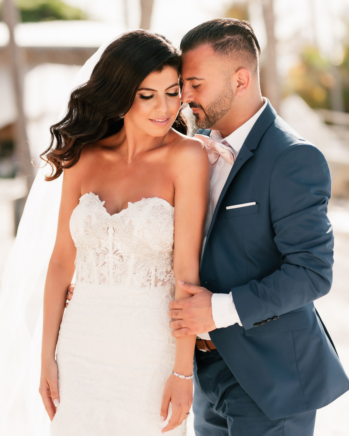 The bride in a white dress and veil, holding the hand, is hugged by the groom. His dark hair and the groom's dark-colored suit stand out vividly against the background of the bride's snow-white dress.