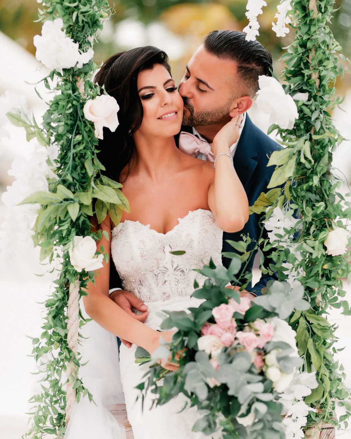 The groom embraces the bride at the altar decorated with white flowers and green leaves. The bride is holding a bouquet of flowers.