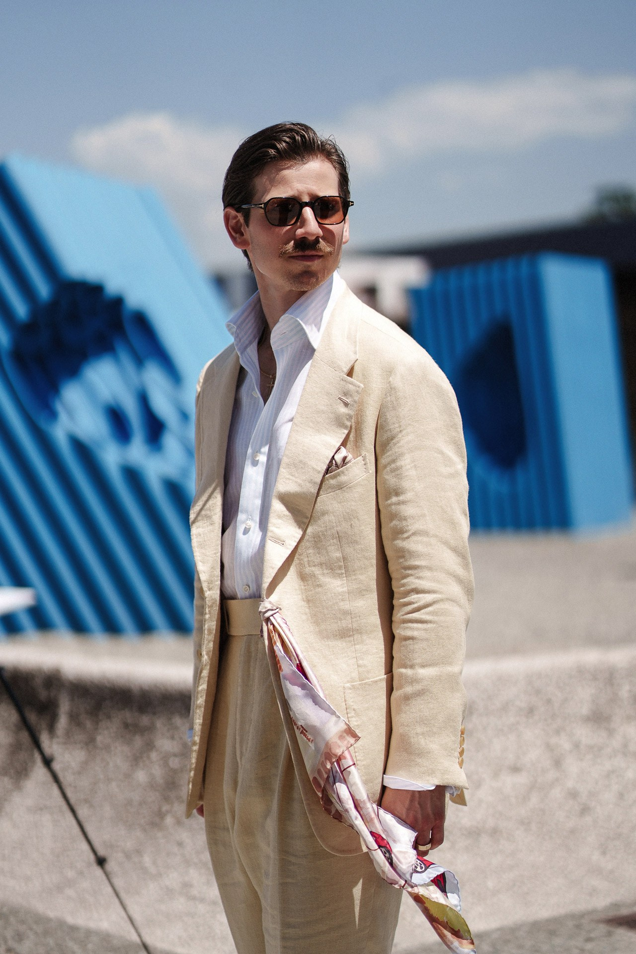 Ritratto street style da Pitti Uomo 110, ospite in completo estivo crema con foulard fantasia vicino all’installazione blu