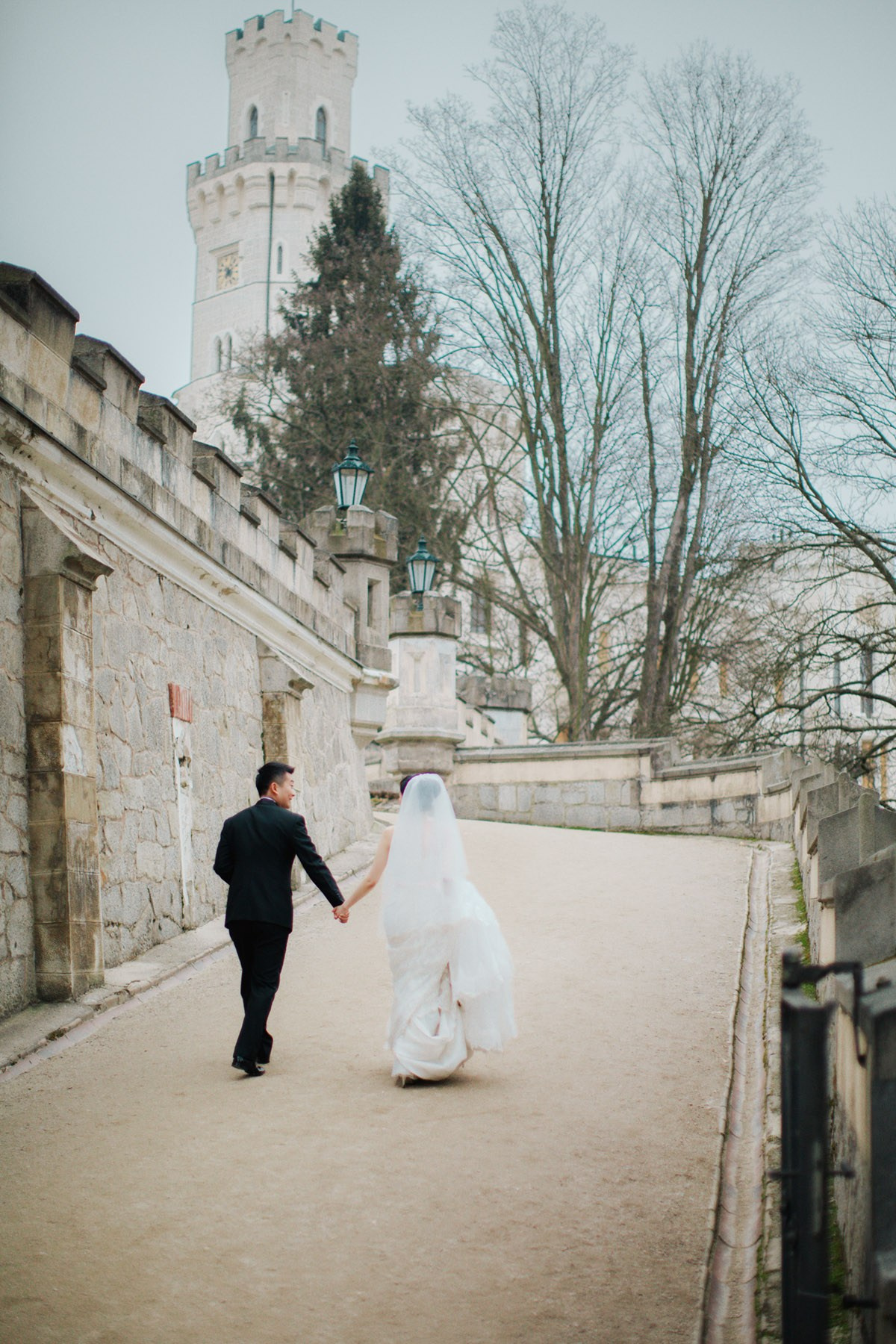 A groom takes his brides hand as they walk up a rampart towards the impressive State Chateau of Huboka during their winter destination wedding in Bohemia.