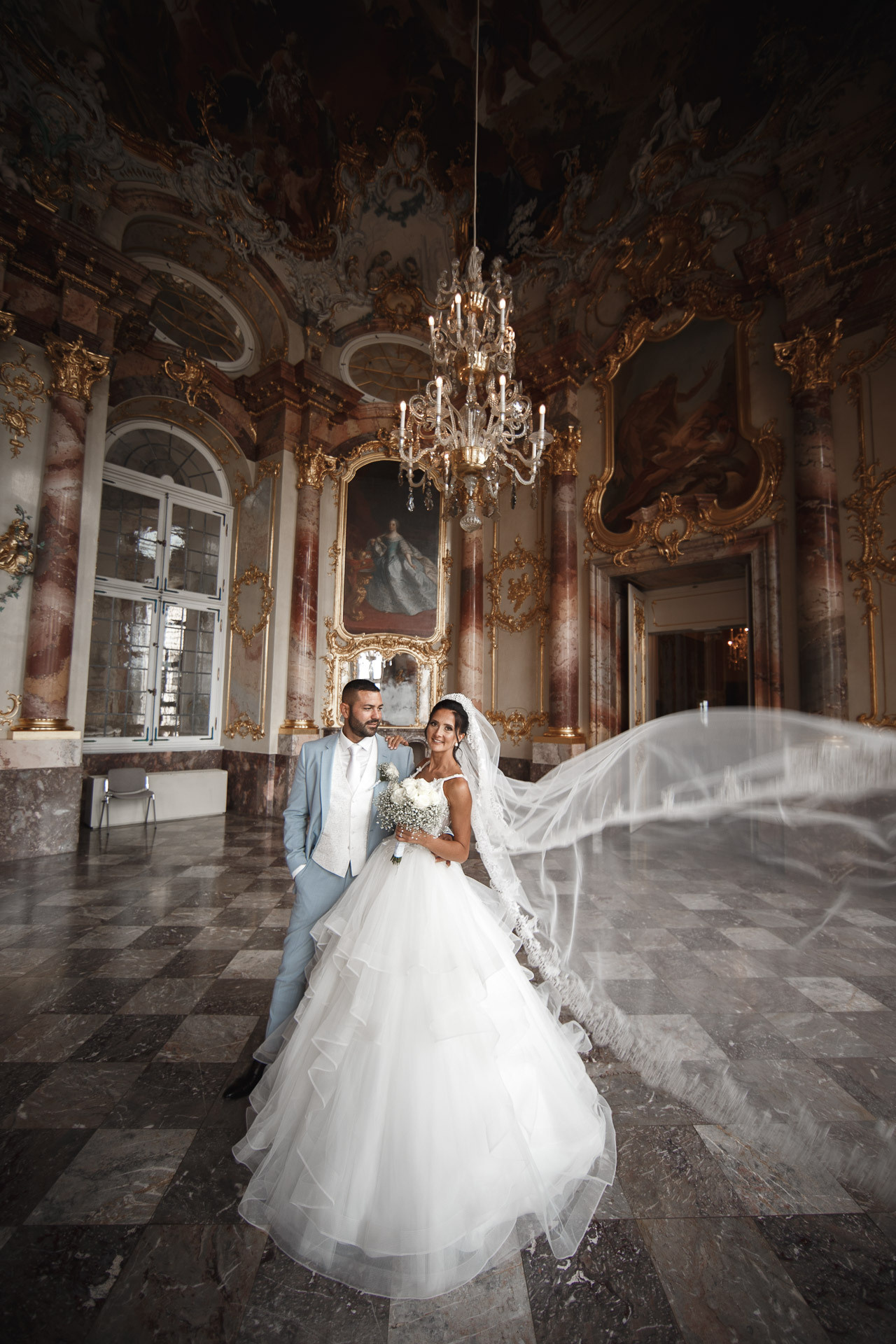 Die Braut und der Brätigam posieren vor einem Kronleuchter im Schloss und strahlen vor Eleganz.