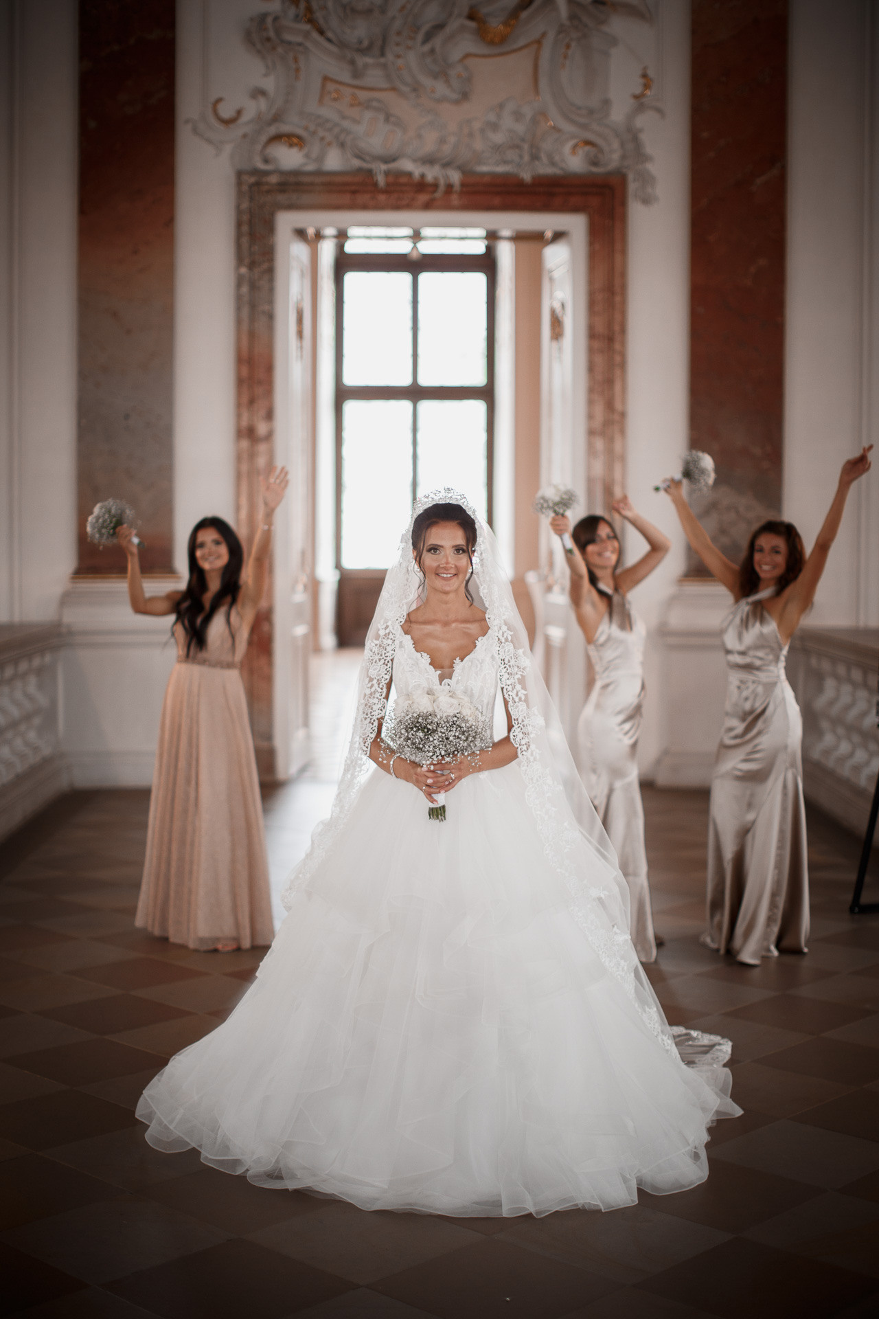 Die Braut und ihre Brautjungfern halten liebevoll Blumen in den Händen und lächeln vor dem Schlosspanorama.