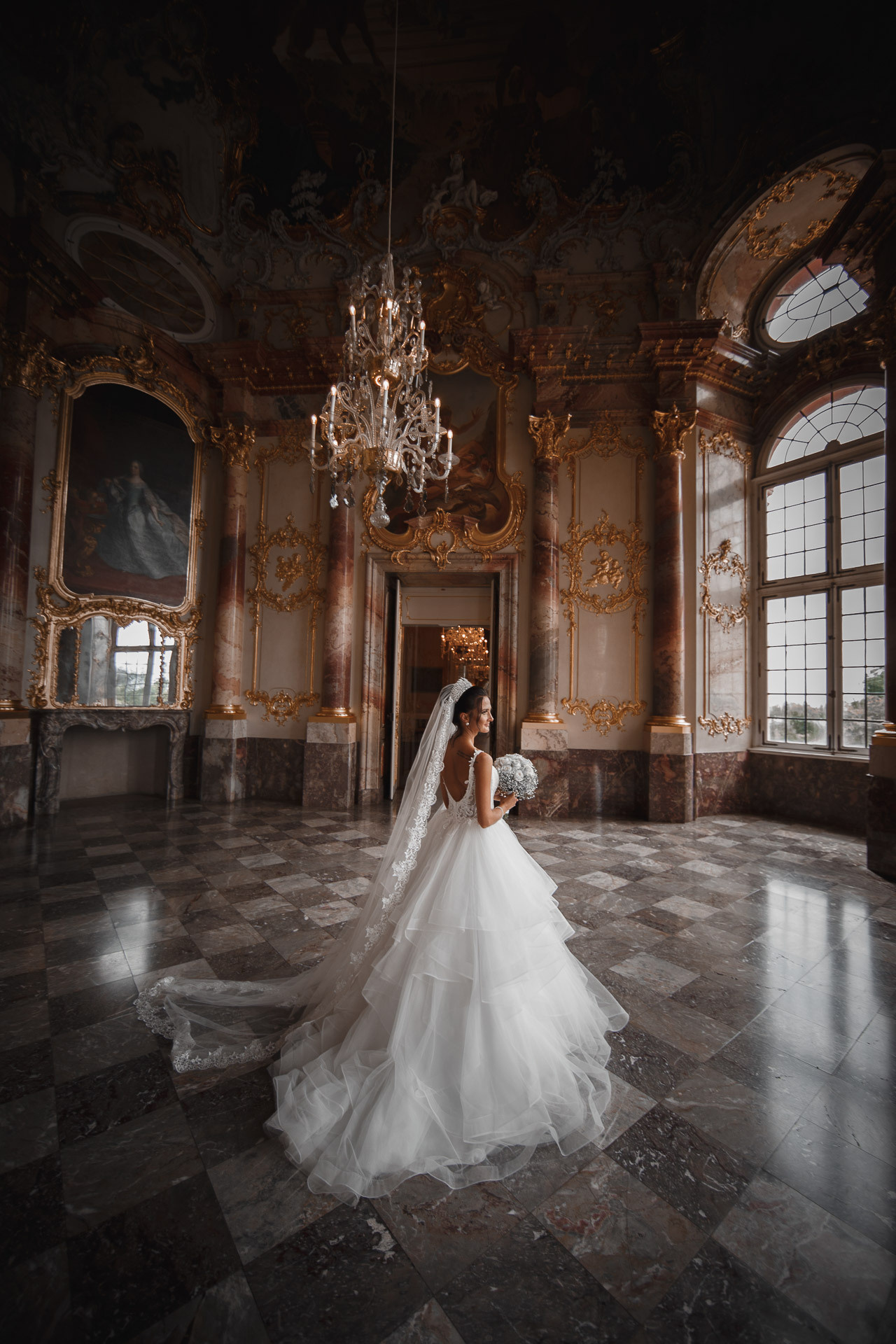 Die Braut posiert vor einem Kronleuchter im Schloss und strahlt vor Eleganz.
