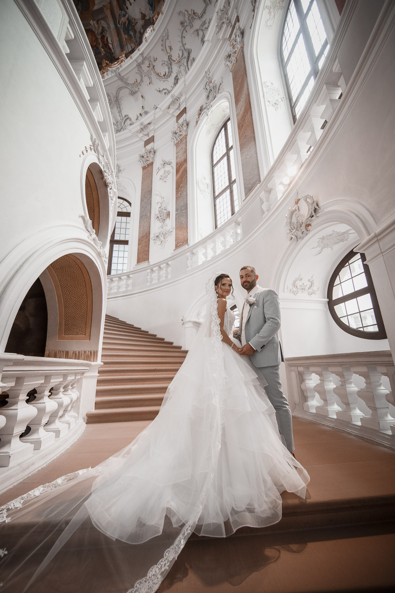 Das Brautpaar lächelt fröhlich, während es Hand in Hand auf der historischen Treppe im Schloss Bruchsal steht.