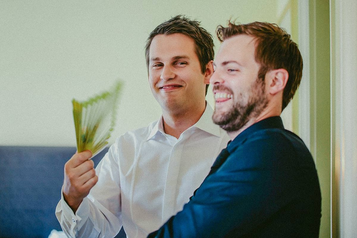 Groom Dan is using a fan to cool his smiling best man in the bridal suite