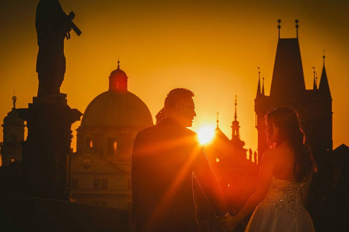 Bride and groom are silhouetted against the medieval landscape of Prague atop the Charles Bridge at sunrise.
