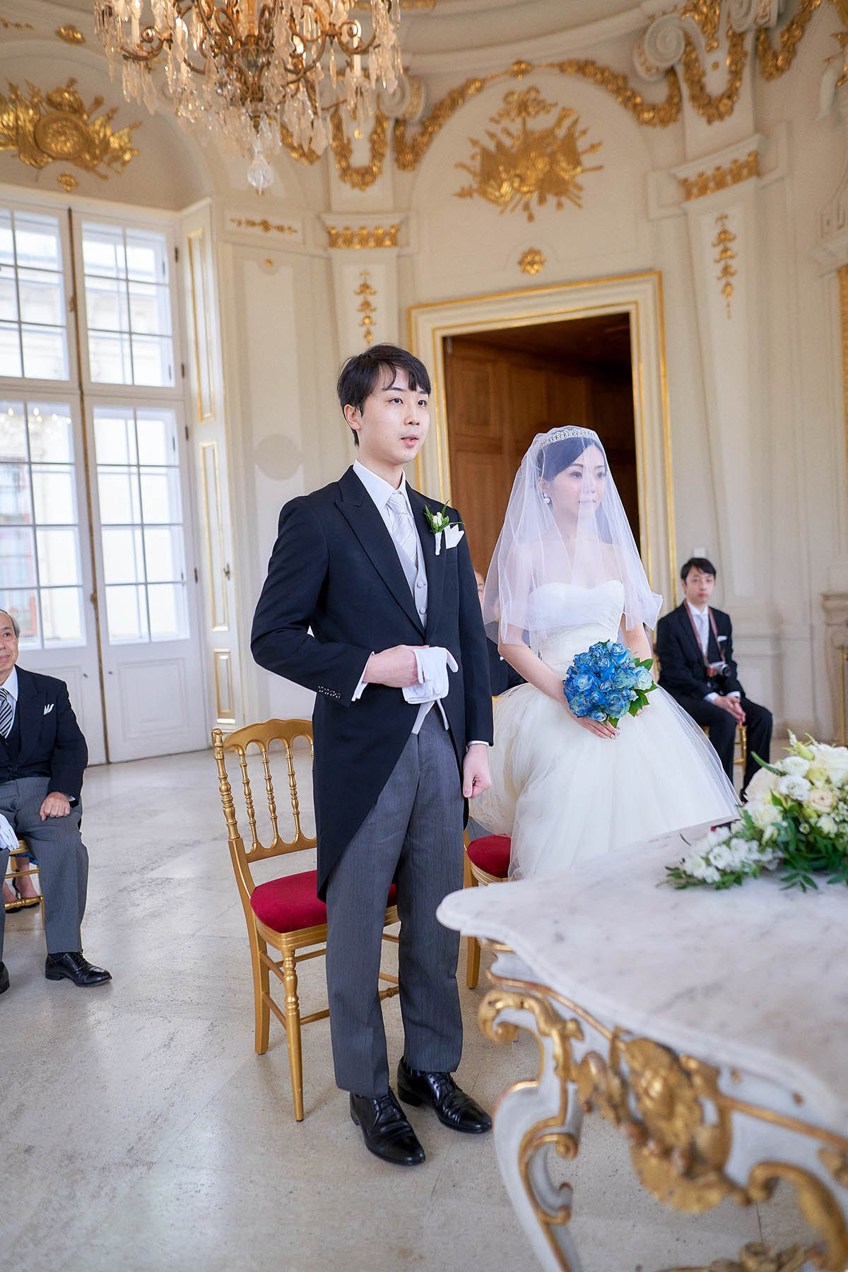 Katsuya and Hitomi stand near the ceremonial table as they are welcomed to the Belvedere Palace by the officiant.