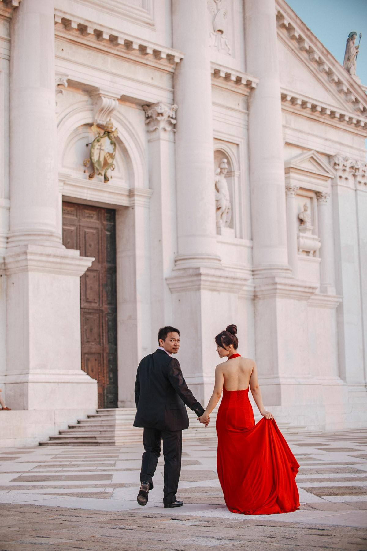 A Thai woman wearing a bold red dress holds hands with her fiancee as they explore a remote island in the lagoons of Venice during the Golden Light Hour.
