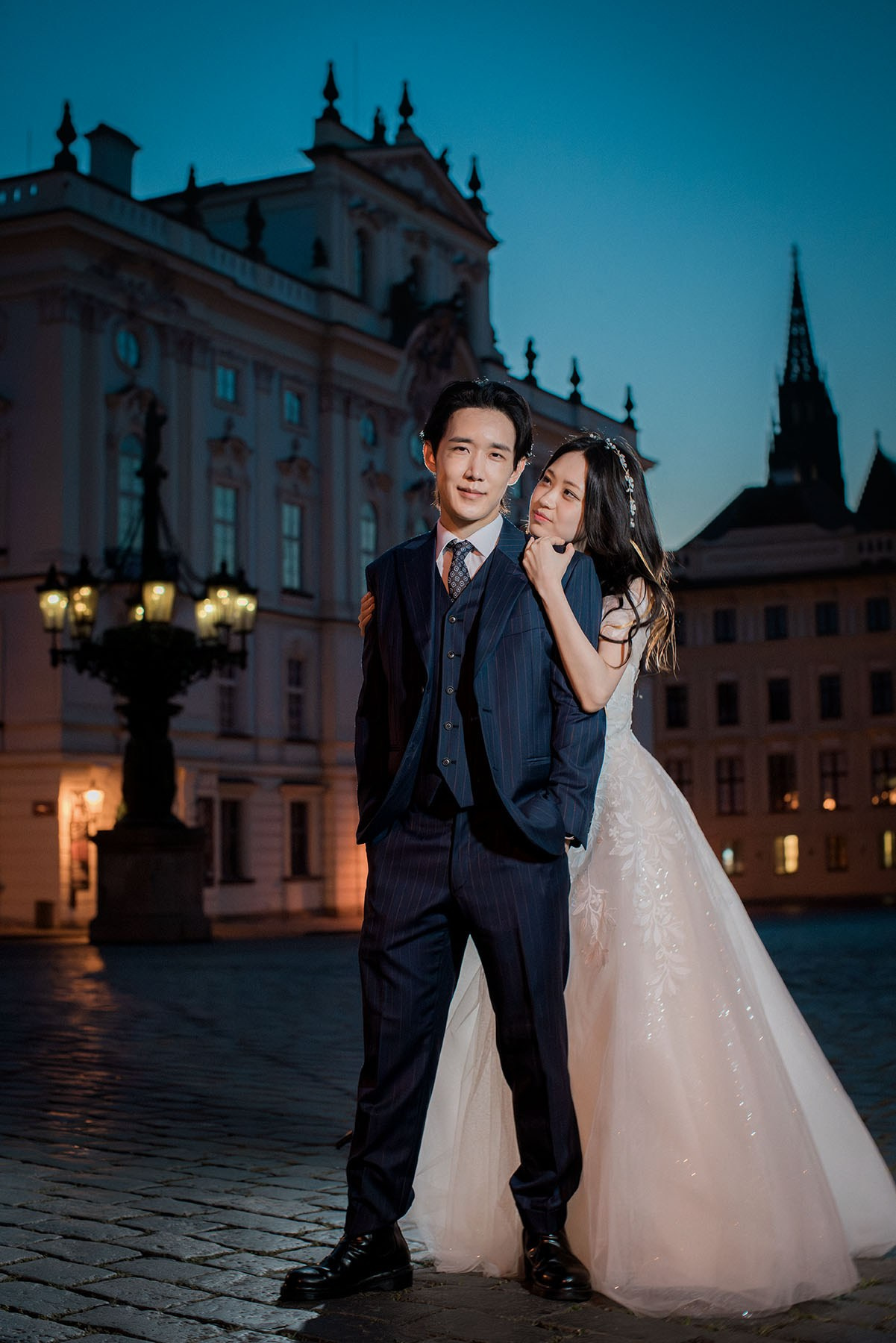 A sexy, good-looking Korean bride stands behind her groom as they pose for a photo at Prague Castle at night.