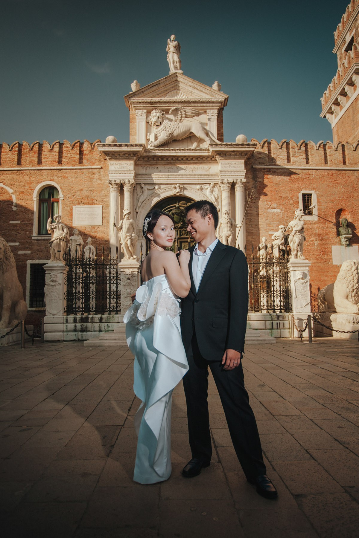 A Thai bride wearing a traditional white wedding dress poses with her fiancee in front of the historic Arsenale fortress in historic Venice during their destination Love Story photo session.