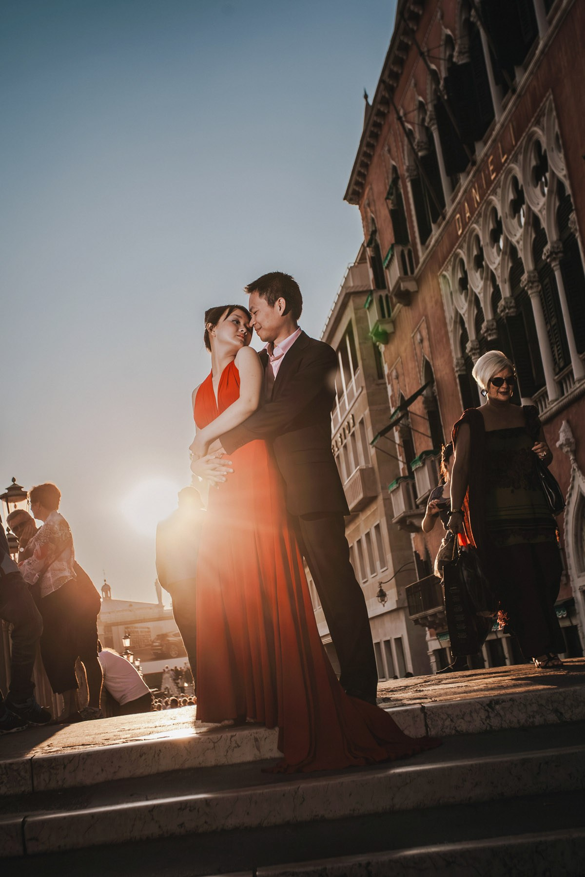 As the late afternoon sunlight flares around them, a Thai woman wearing a sexy red dress is embraced by her fiancee at a bridge near the Hotel Danieli in Venice as an elegantly dressed woman walks by.