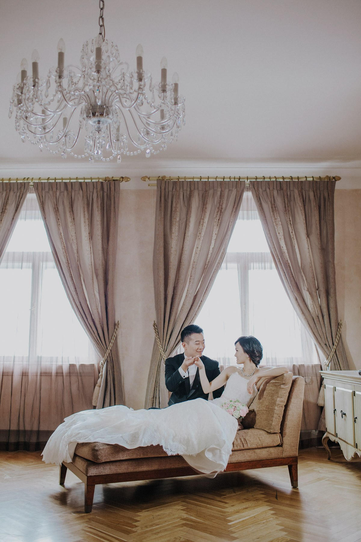An antique sofa is the perfect setting for the reclining Hong Kong bride as her groom takes her hand as he kneels down in their luxury suite in Prague.