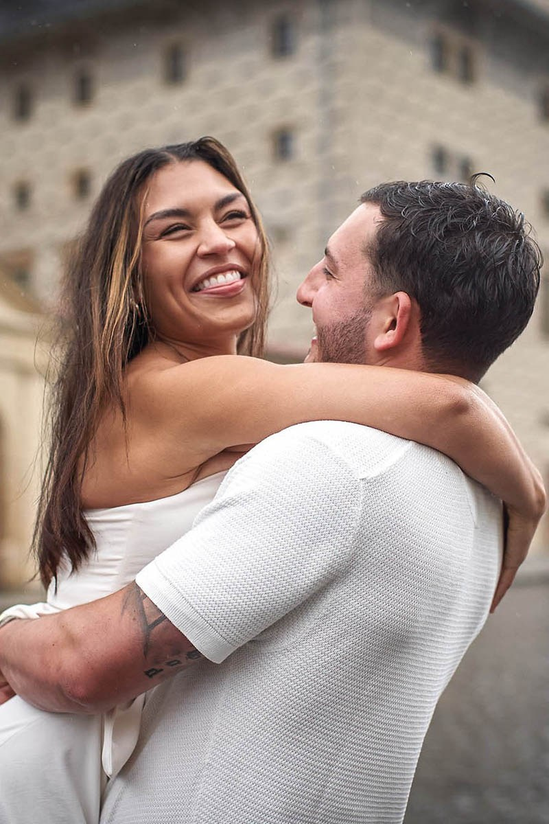 A smiling woman is held aloft by her partner as they stand in the rain at Prague Castle.