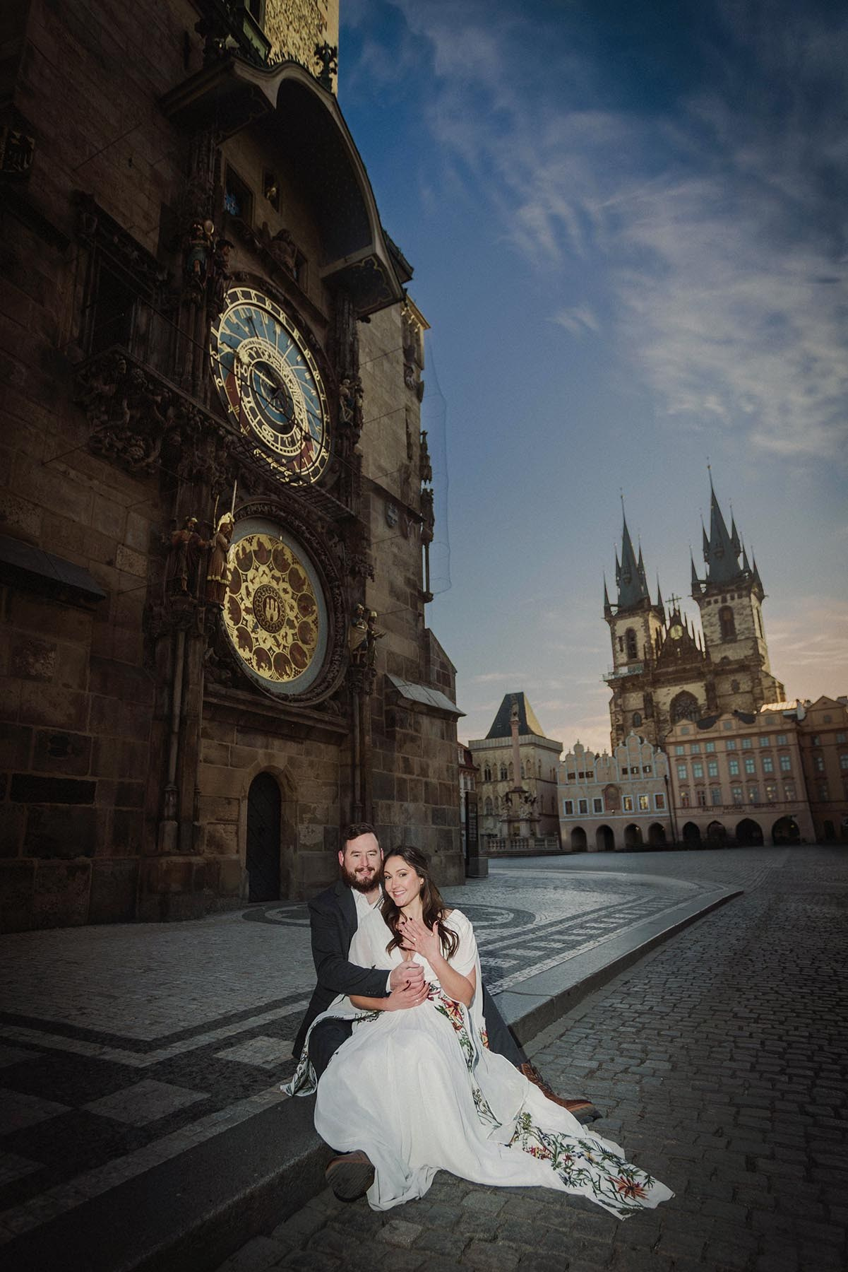 An American couple sitting under the Astronomical Clock as a woman shows off her diamond engagement ring.