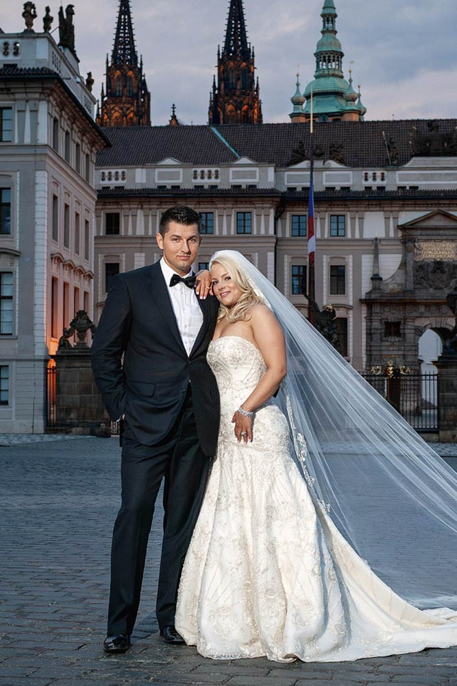 A smiling, confident bride and her groom look towards the camera at Prague Castle at dusk.