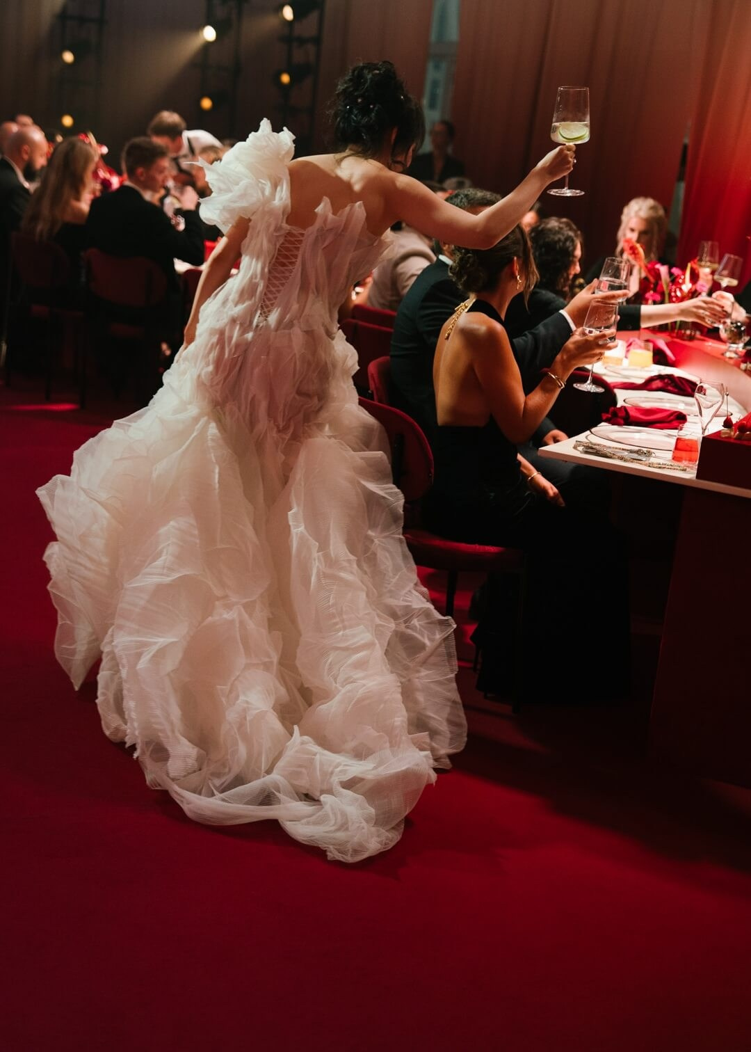 Bride in a dramatic ruffled gown raises her glass to celebrate at a richly lit wedding reception