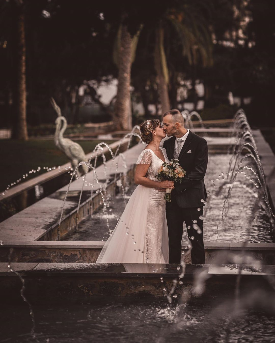 Une mariée et un marié s'embrassant devant une fontaine.