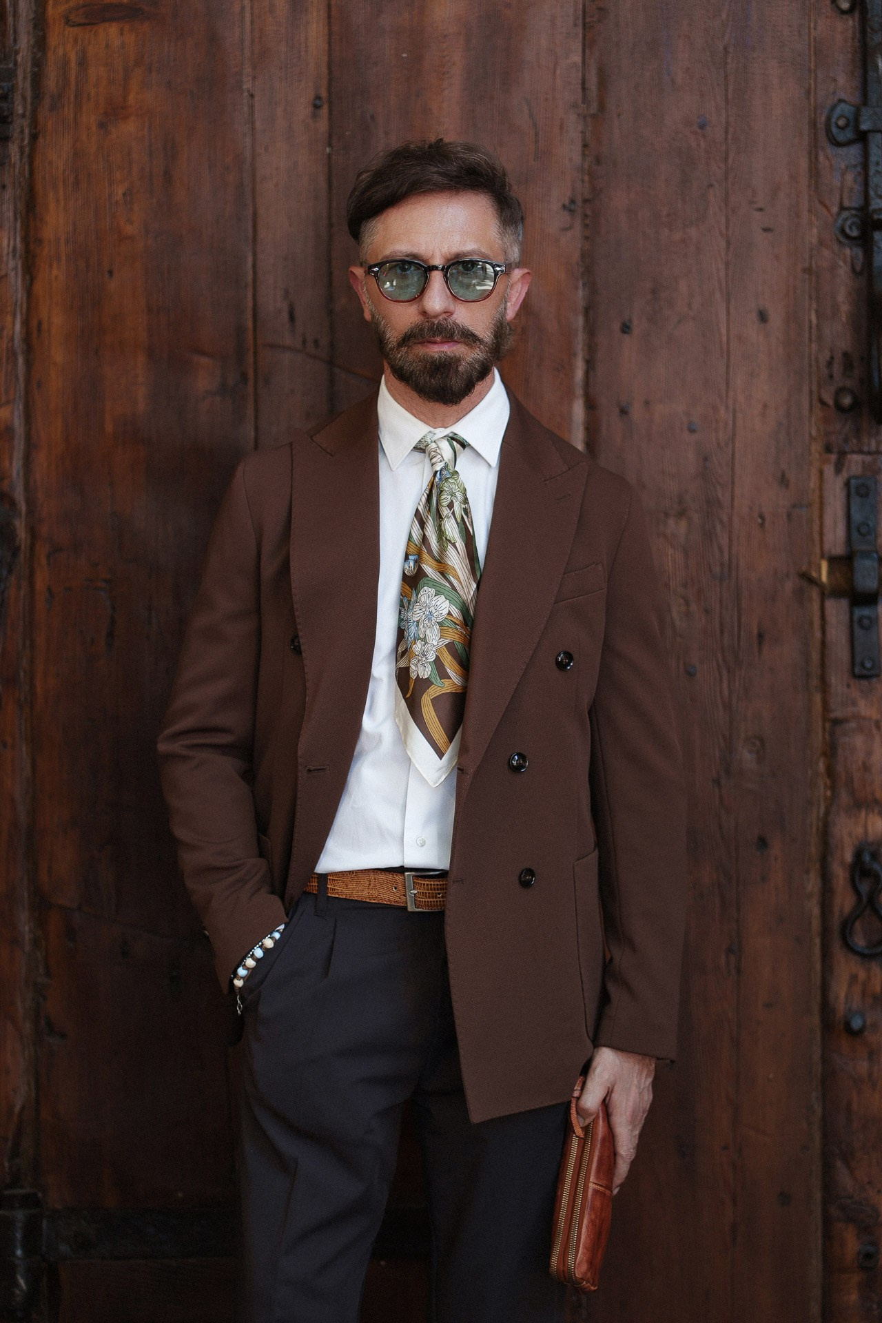 Ritratto menswear da Pitti Uomo 110, ospite in giacca doppiopetto marrone e cravatta fantasia davanti a porte in legno